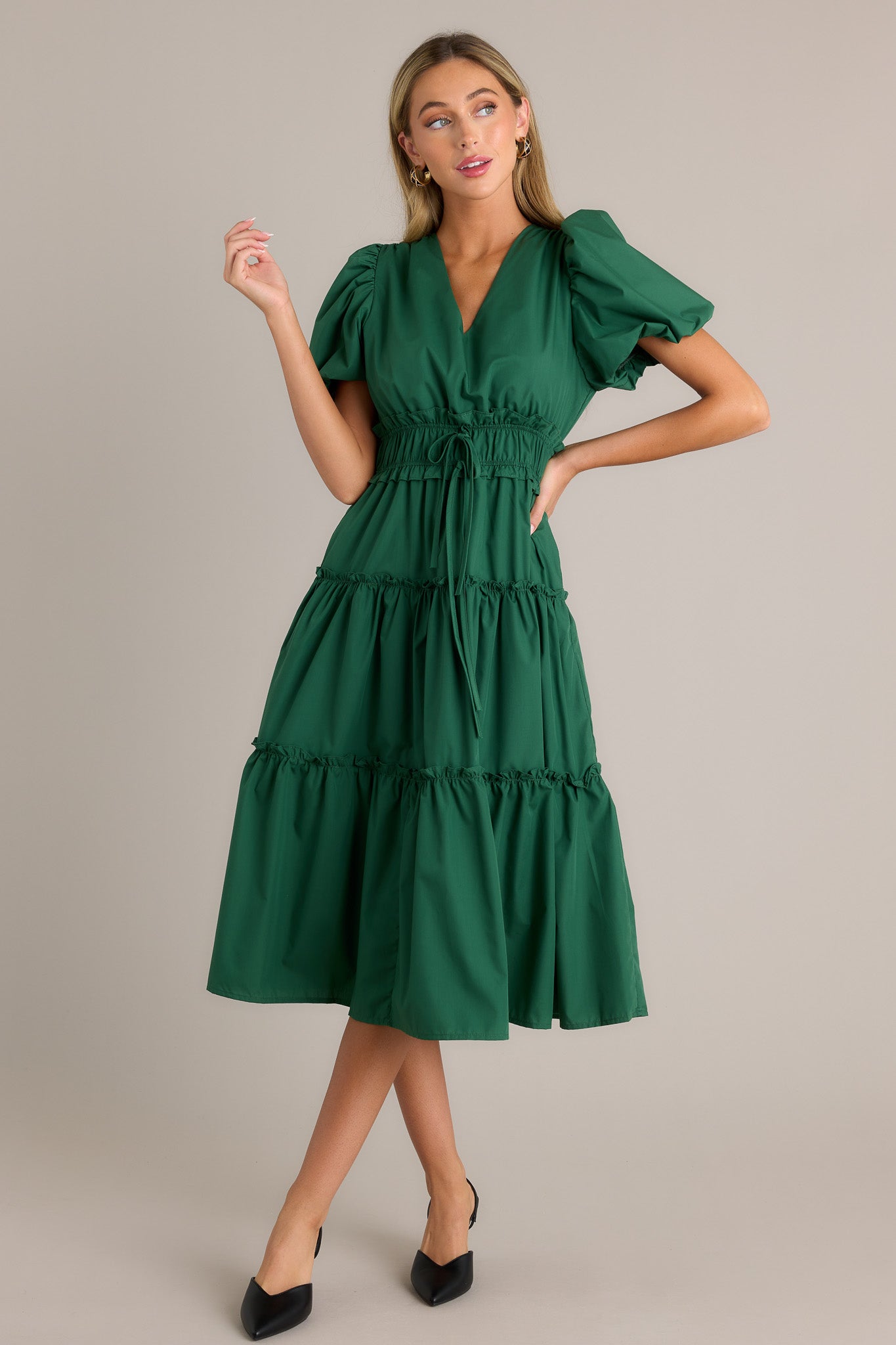 Side view of a green midi dress showcasing the v-neckline, ruffled elastic waistband, self-tie waist features, functional hip pockets, tiered design, and short puffed sleeves.