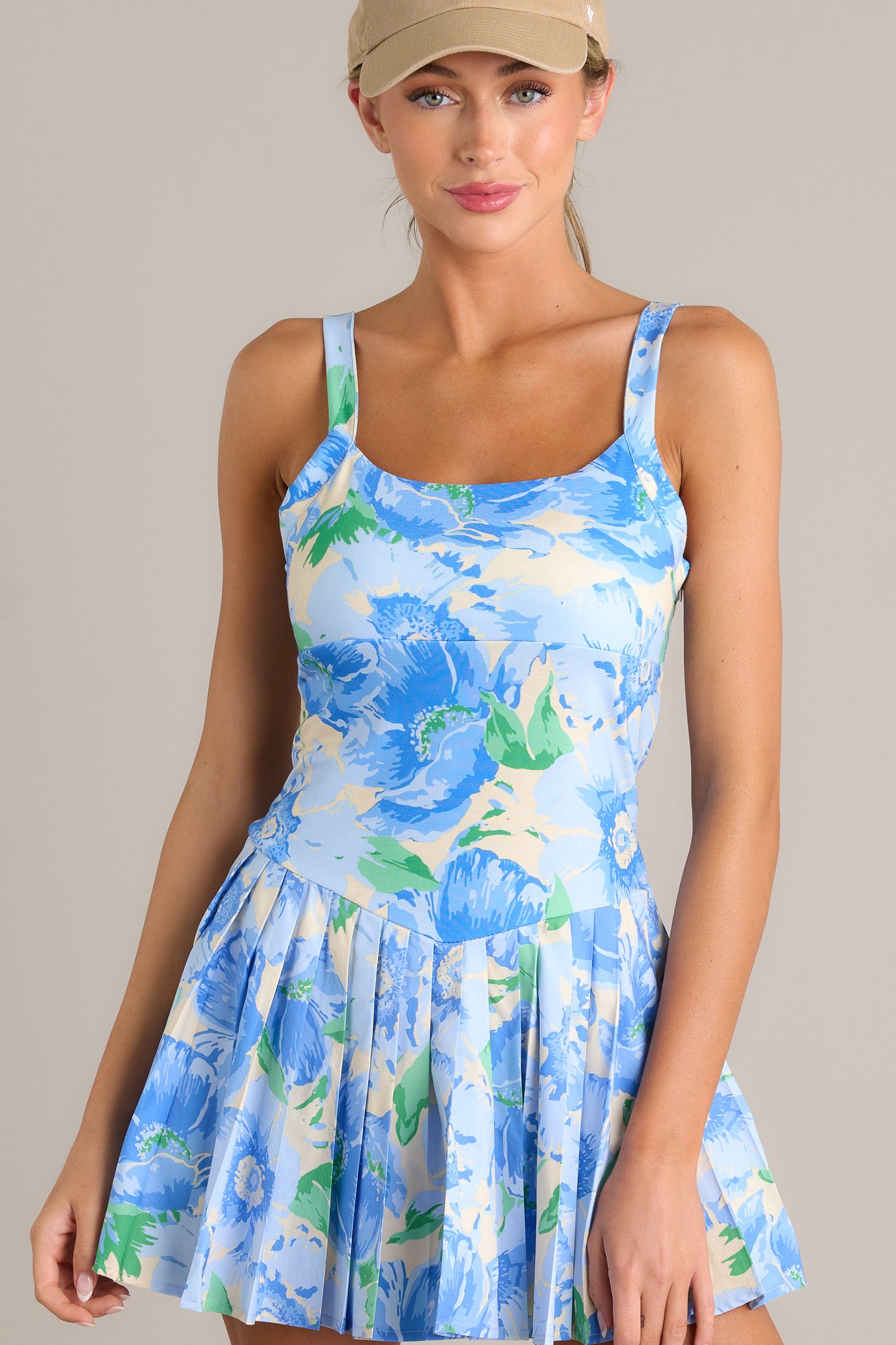 Front view of a blue mini dress featuring a square neckline, thin straps, a discrete side zipper, a floral pattern, and a pleated skirt with built-in shorts underneath.