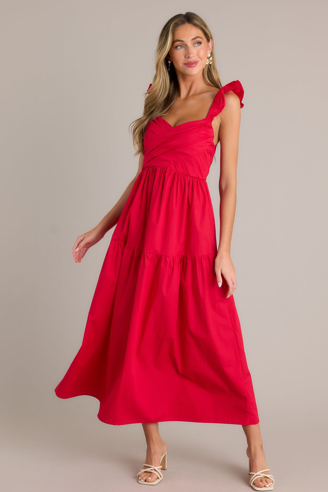 Front angled view of a red midi dress featuring a v-neckline, a pleated bodice, a fully smocked back, a tiered design, layered flutter sleeves, and a flowing silhouette