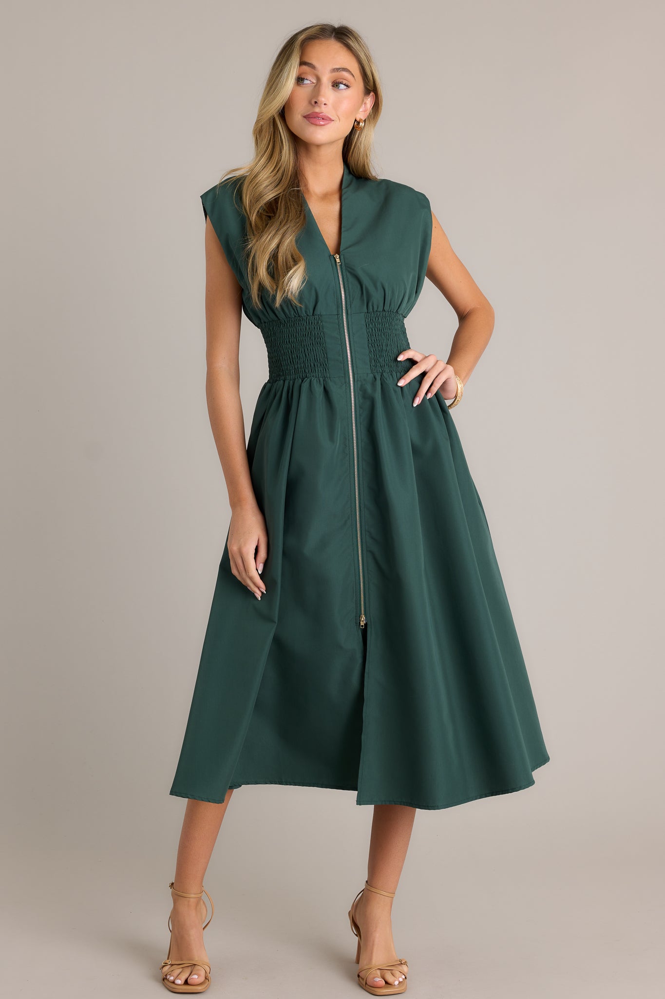 Full length view of a hunter green midi dress with a v-neckline, padded shoulders, a full zipper front, a fully smocked waist, functional hip pockets, and a front slit