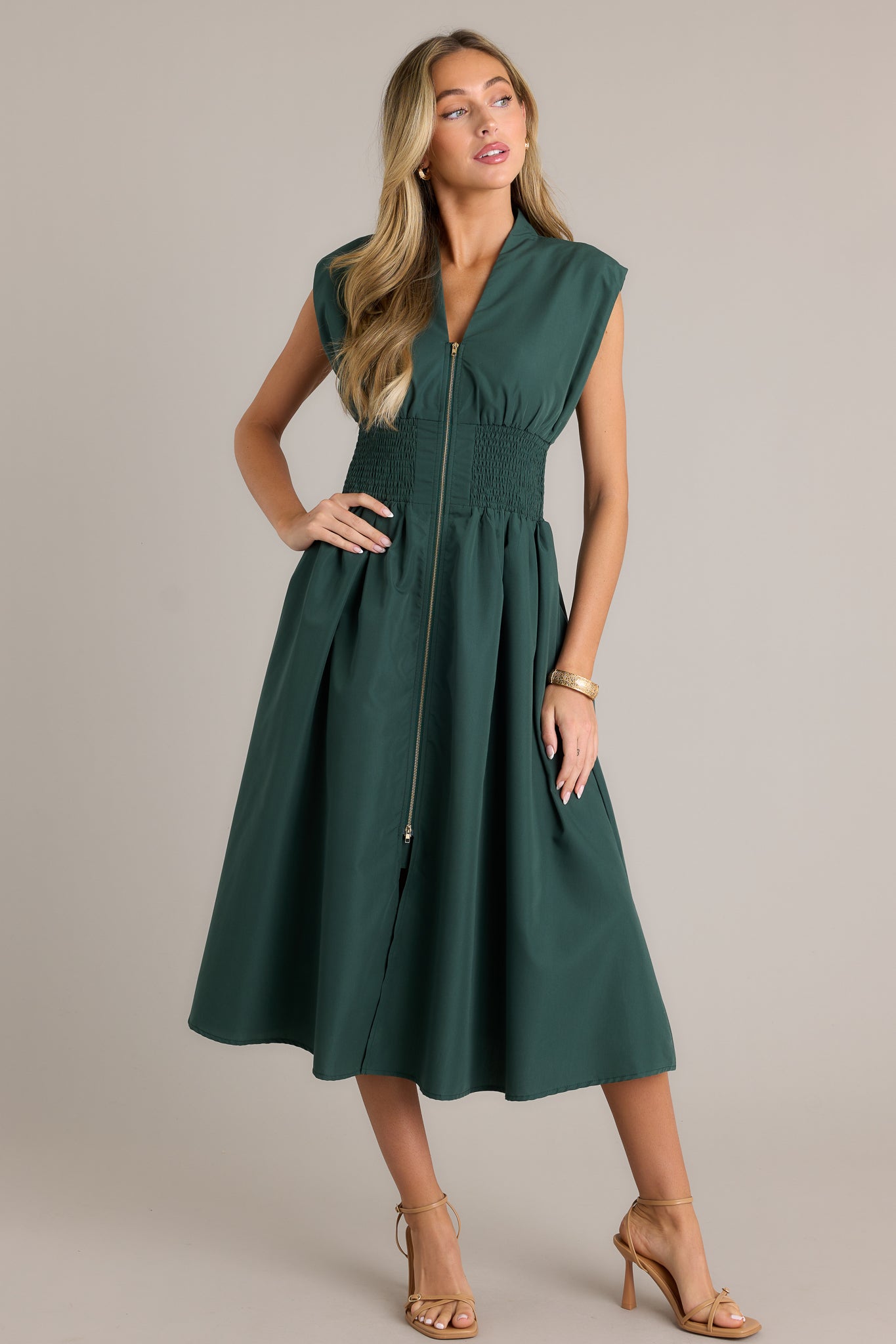 Front angled view of a hunter green midi dress featuring a v-neckline, padded shoulders, a full zipper front, a fully smocked waist, functional hip pockets, and a front slit