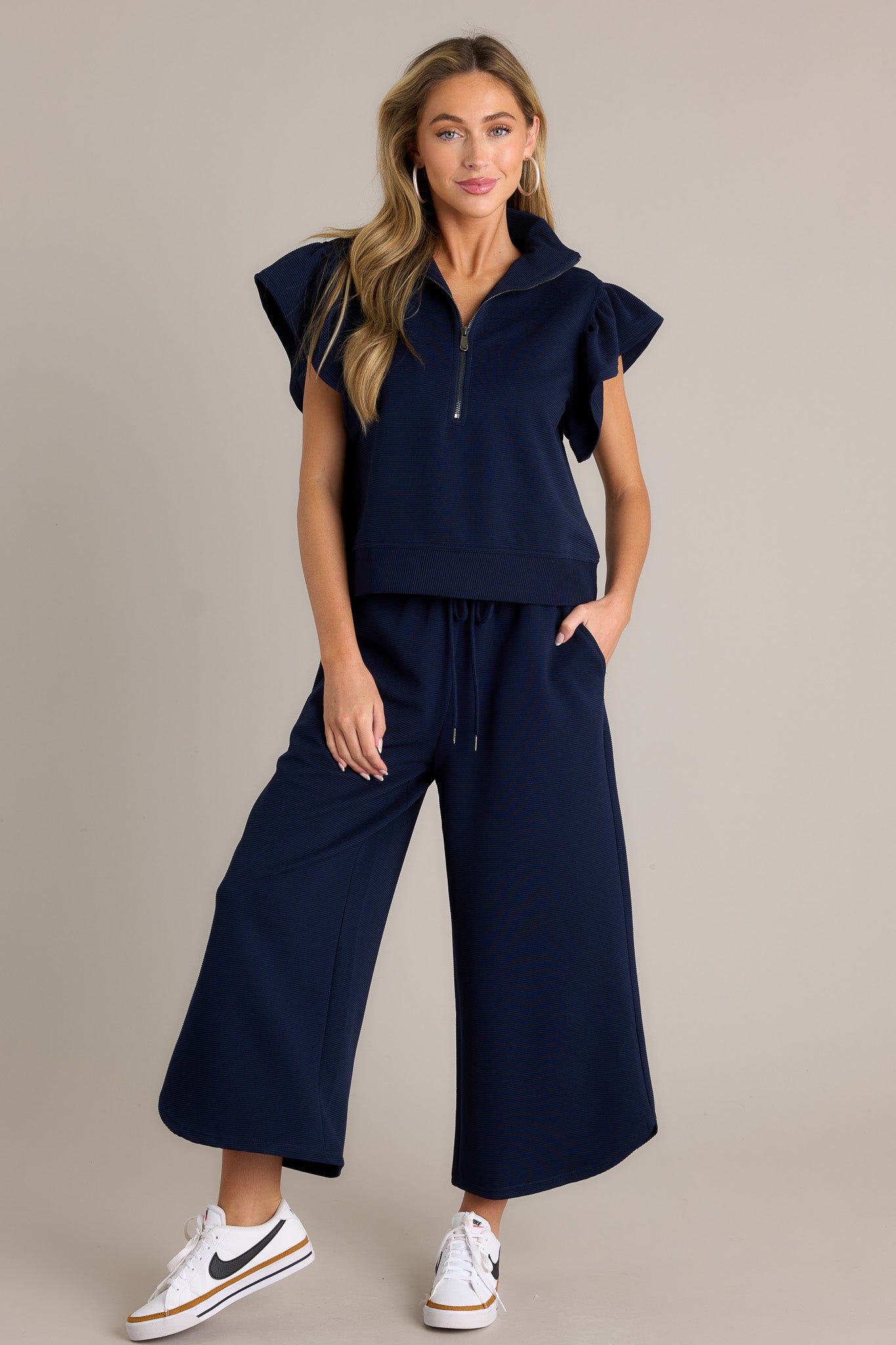 Front angled view of a navy top featuring a functional zip up neckline, a ribbed fabric, wide ruffled sleeves, and a thick hemline