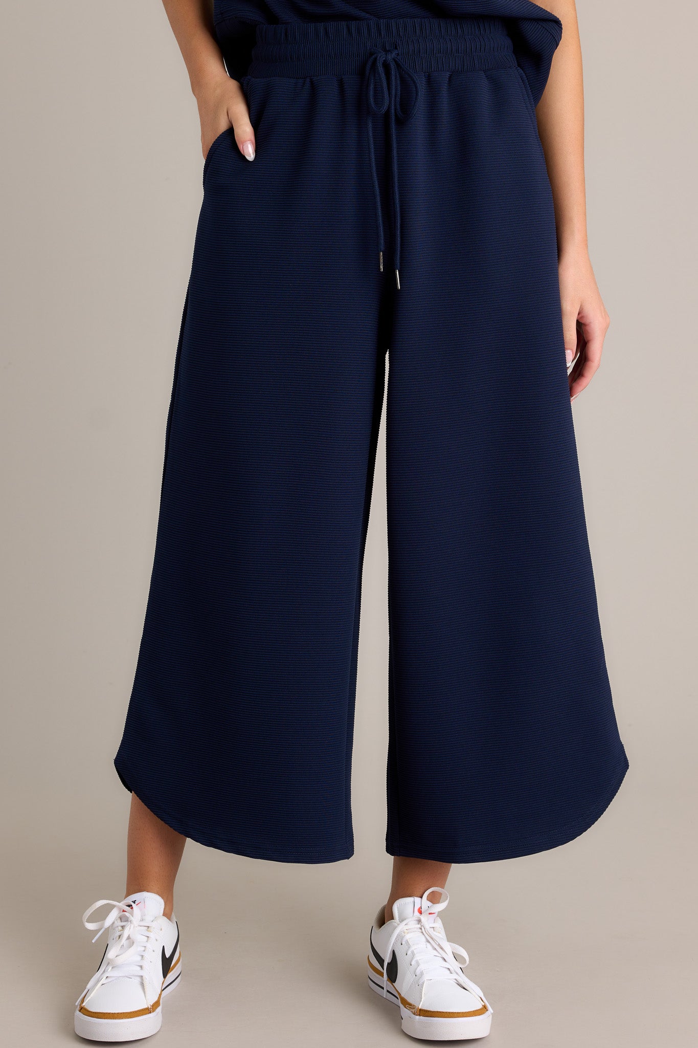 Front angled view of navy pants featuring a high waisted design, an elastic waistband, a self-tie drawstring, functional hip pockets, a ribbed texture, a wide leg, and a scooped hemline