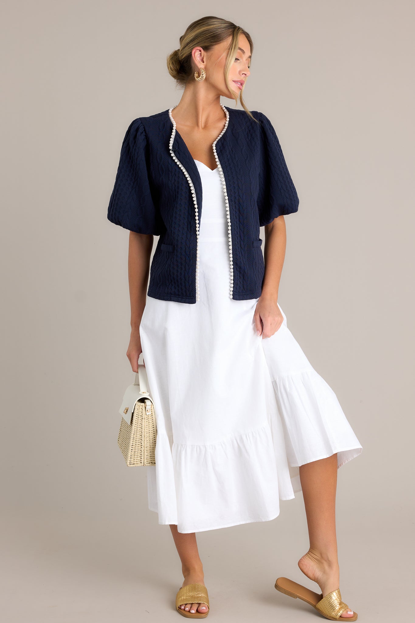 Full length view of a navy short sleeve jacket with a v-neckline, a hook & bar closure, faux pearl trim, a quilted design, faux pockets, and elastic cuffed puff sleeves