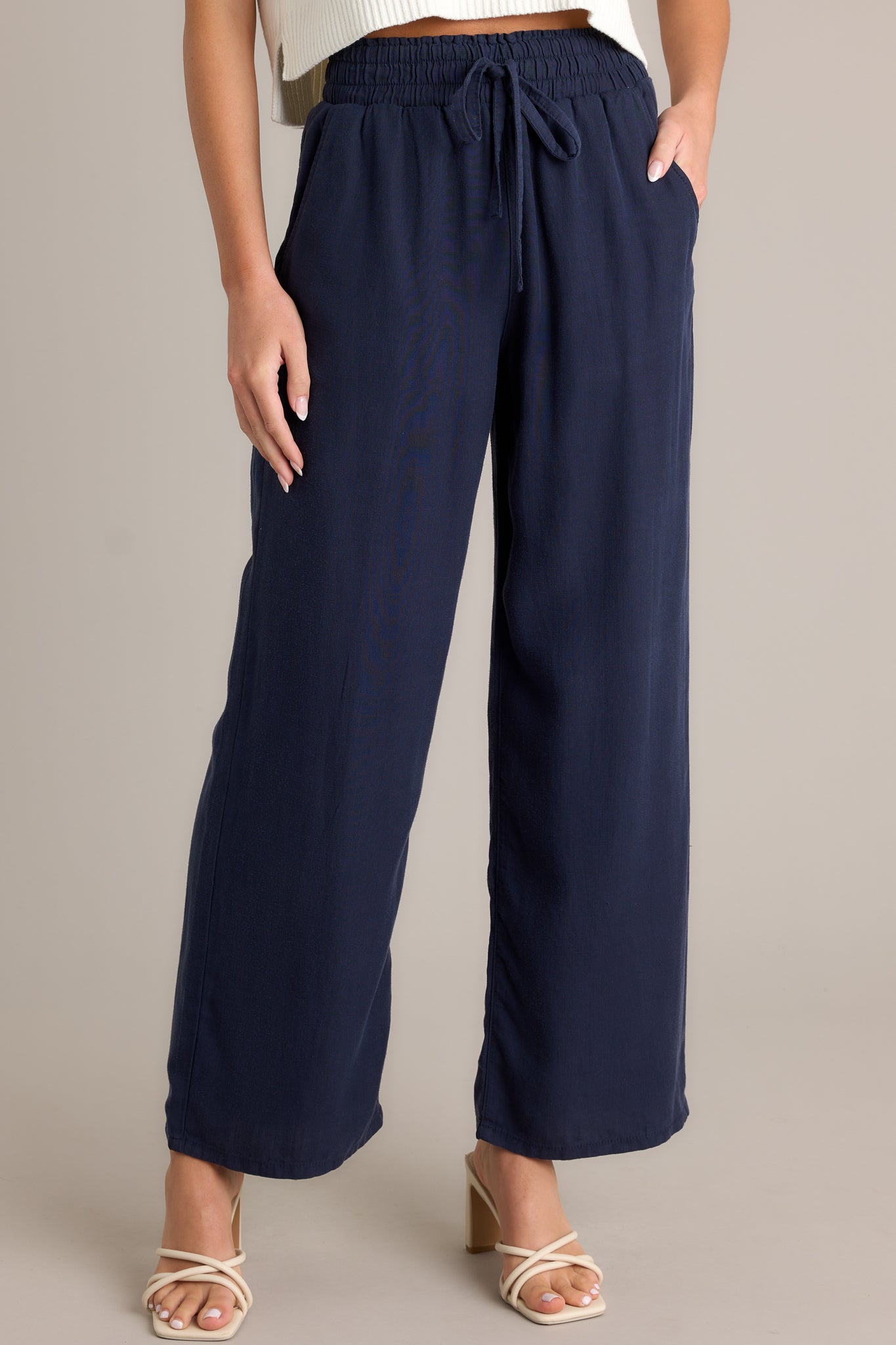 Angled front  view of the material of these navy pants that feature a high waisted design, an elastic waistband with a self-tie drawstring, functional front & back pockets, and a wide leg.