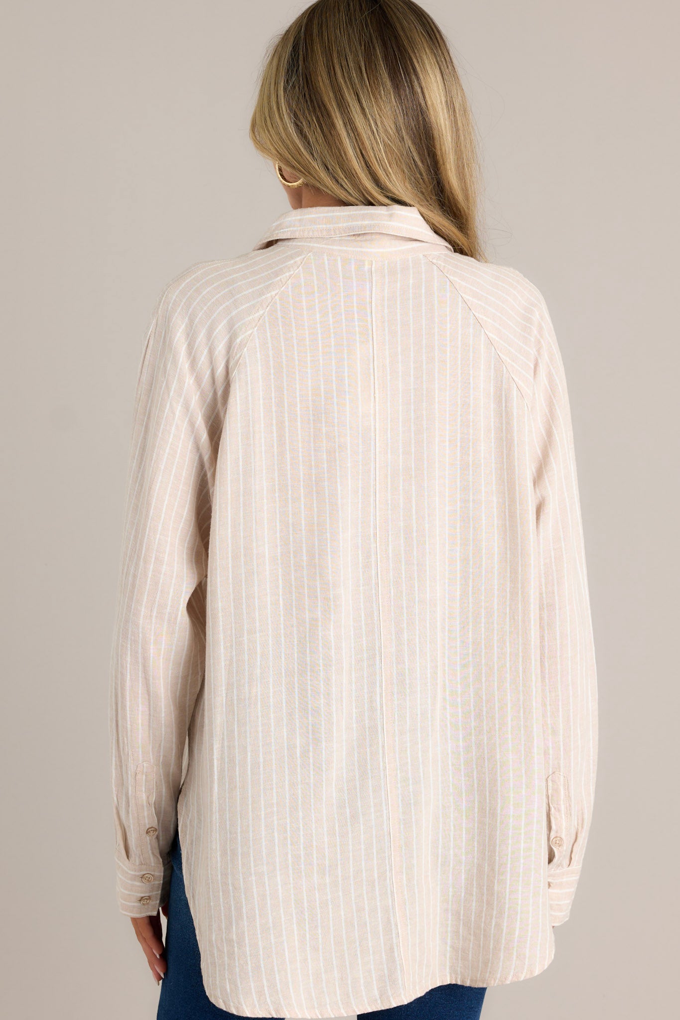 Back view of a beige stripe top highlighting the overall fit, button cuffed long sleeves, and split high low hemline.