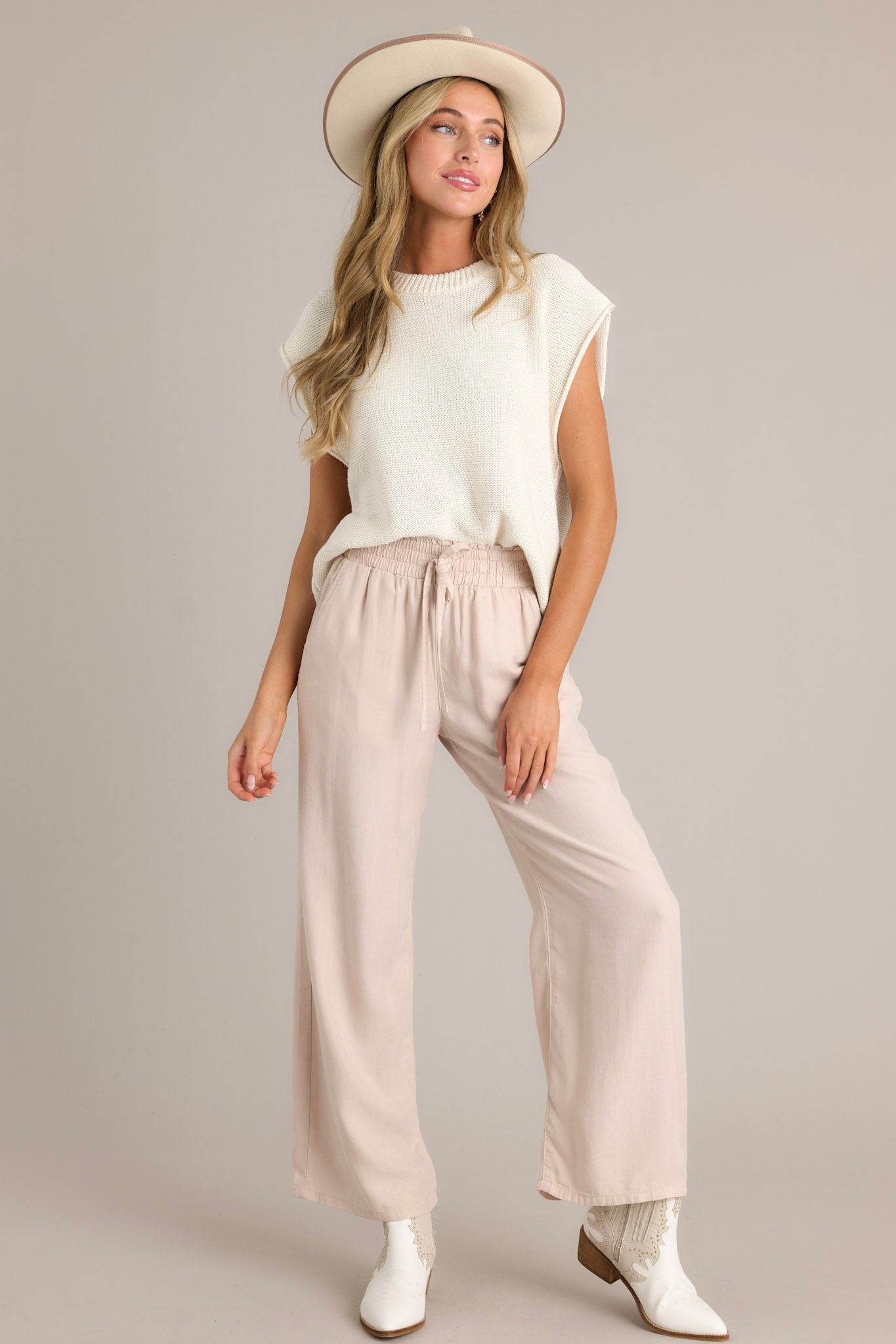 Full length view of beige pants with a high waisted design, an elastic waistband with a self-tie drawstring, functional front & back pockets, and a wide leg