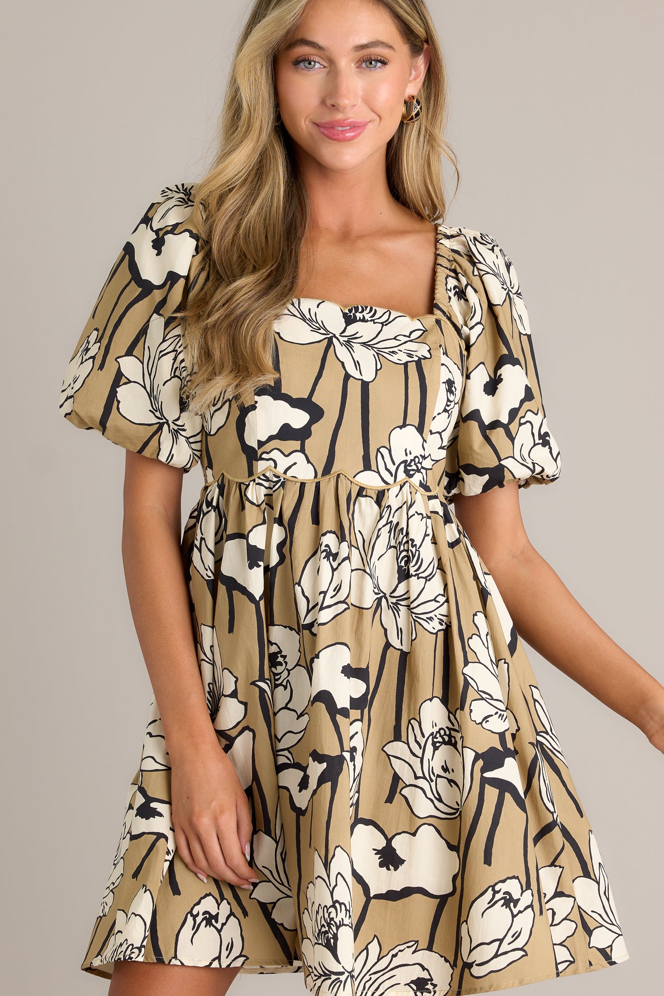 Front view of a taupe mini dress featuring a scalloped neckline, a fully smocked back, a scalloped waistline, a flowing silhouette, and elastic cuffed puff sleeves.