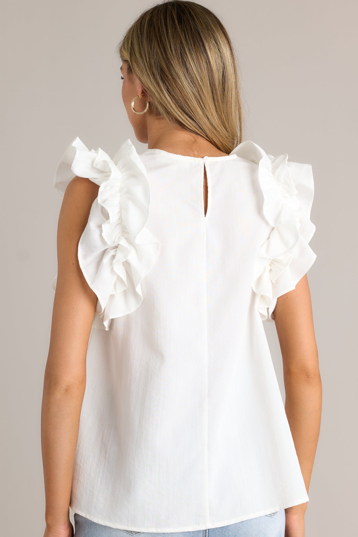 Back view of this ivory top that features a high crew neckline, a keyhole with a button closure at the back of the neck, and ruffled shoulder detailing.