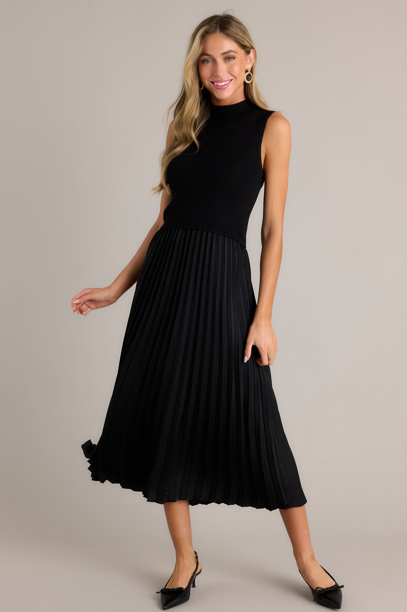 This black sleeveless dress features a high neckline, a ribbed knit bodice, a pleated skirt, a sleeveless design and a flowing silhouette.