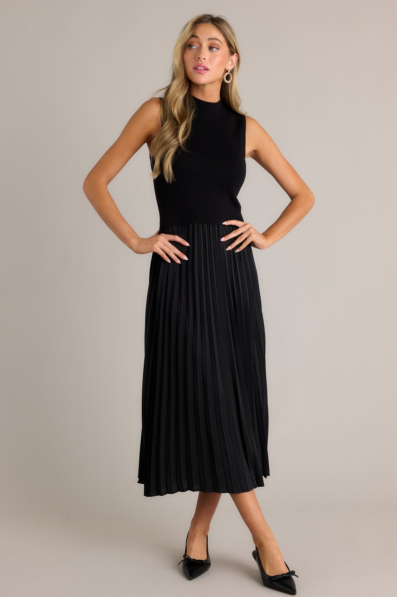 Front view of this black sleeveless dress featuring a high neckline, a ribbed knit bodice, a pleated skirt, a sleeveless design and a flowing silhouette.