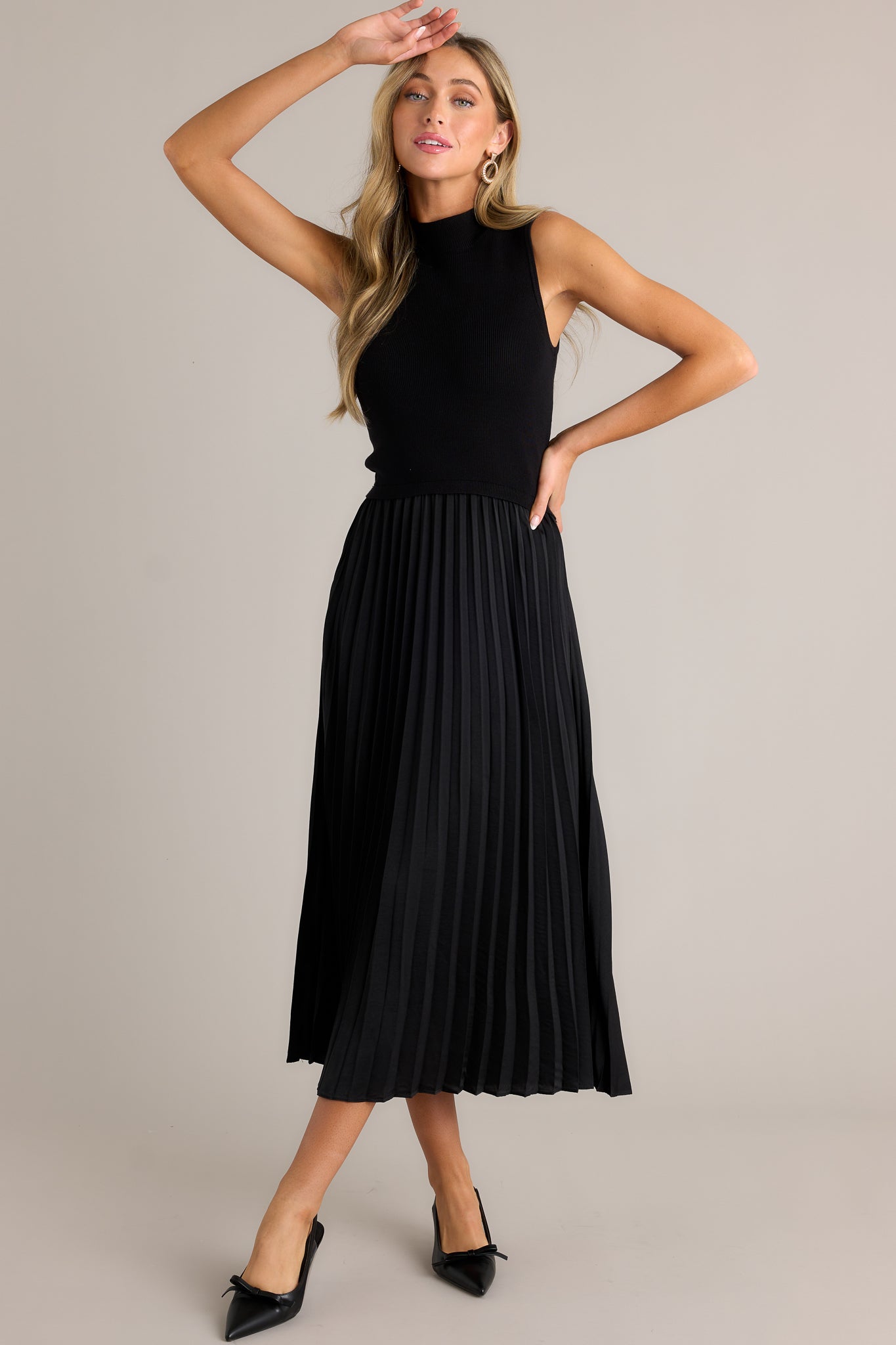 Full body view of this black sleeveless dress featuring a high neckline, a ribbed knit bodice, a pleated skirt, a sleeveless design and a flowing silhouette.