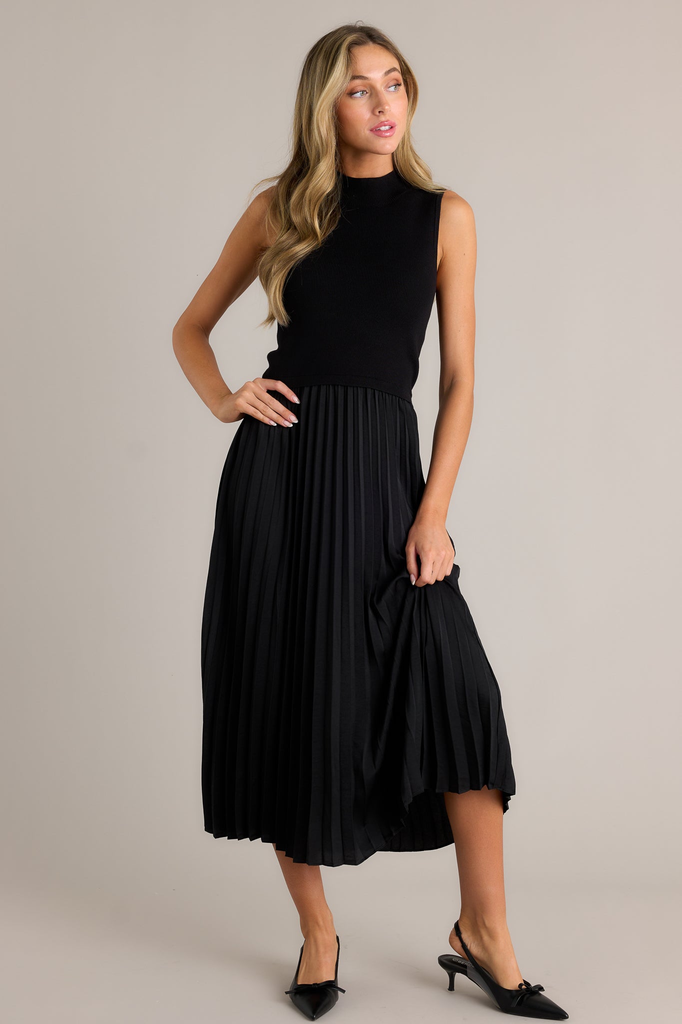 Posed view of this black sleeveless dress featuring a high neckline, a ribbed knit bodice, a pleated skirt, a sleeveless design and a flowing silhouette.