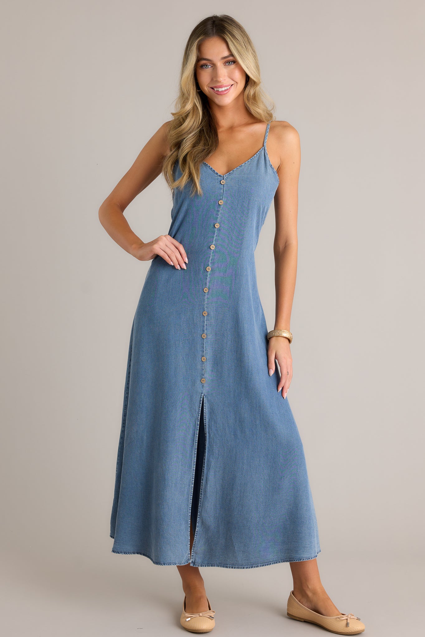 Front angled view of a chambray maxi dress featuring a v-neckline, button front design, smocked back insert, adjustable spaghetti straps, and an open leg slit