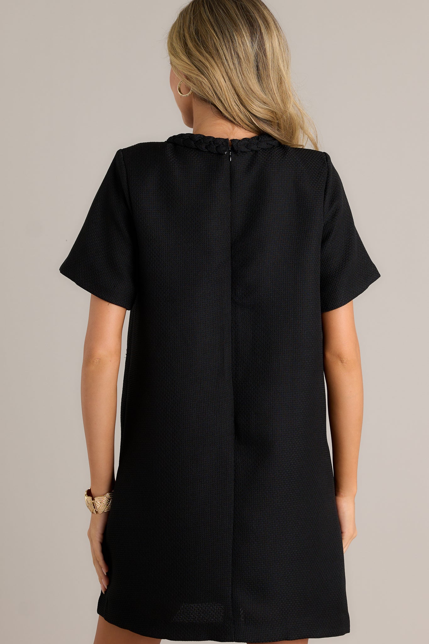 Back view of this black mini dress featuring a braided crew neckline, gold accent buttons, faux hip pockets, a tweed material and short sleeves.