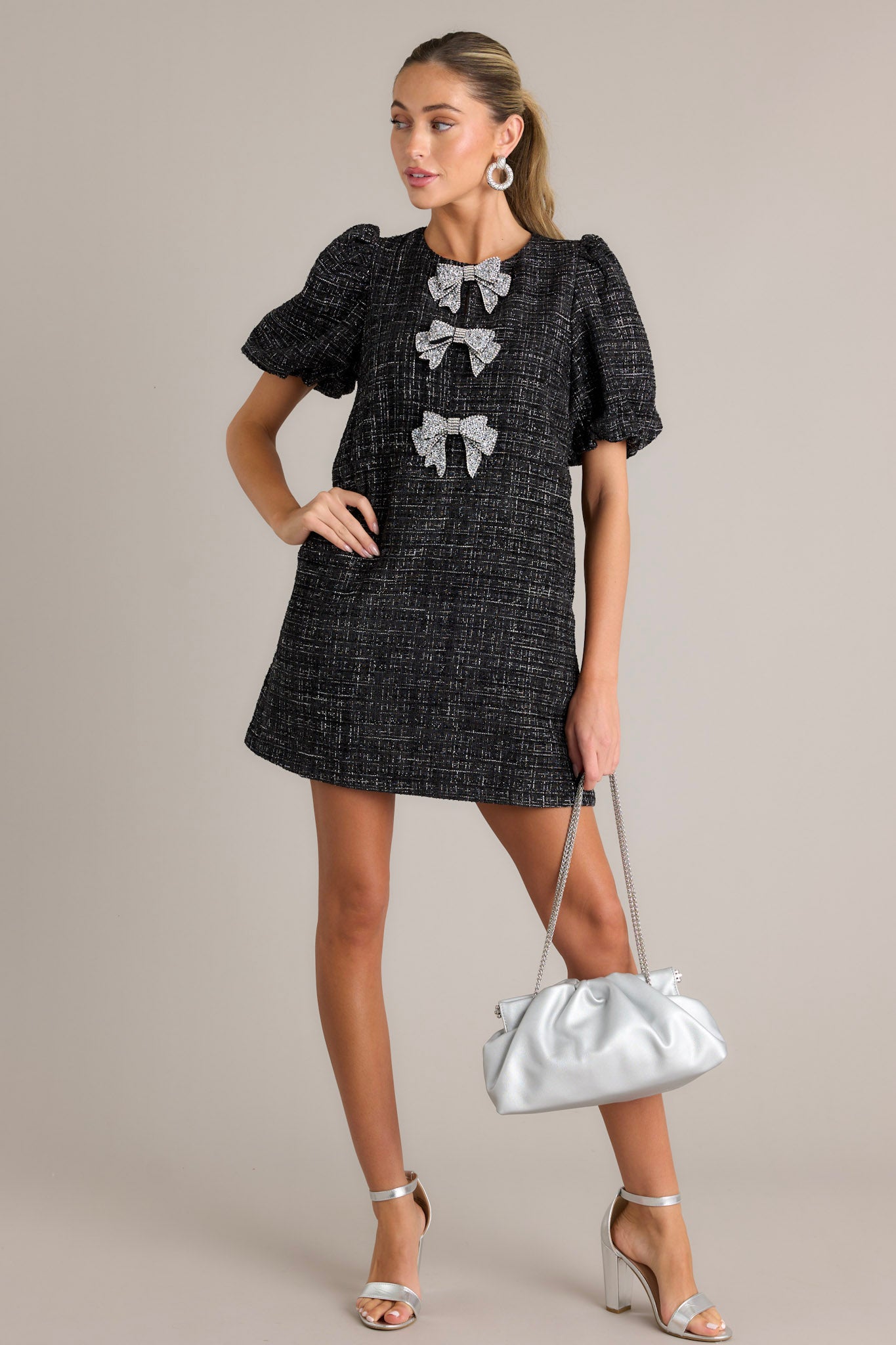  A black tweed dress featuring a round neckline, short puff sleeves, and three large silver glitter bows, accessorized with a silver purse and silver heels.