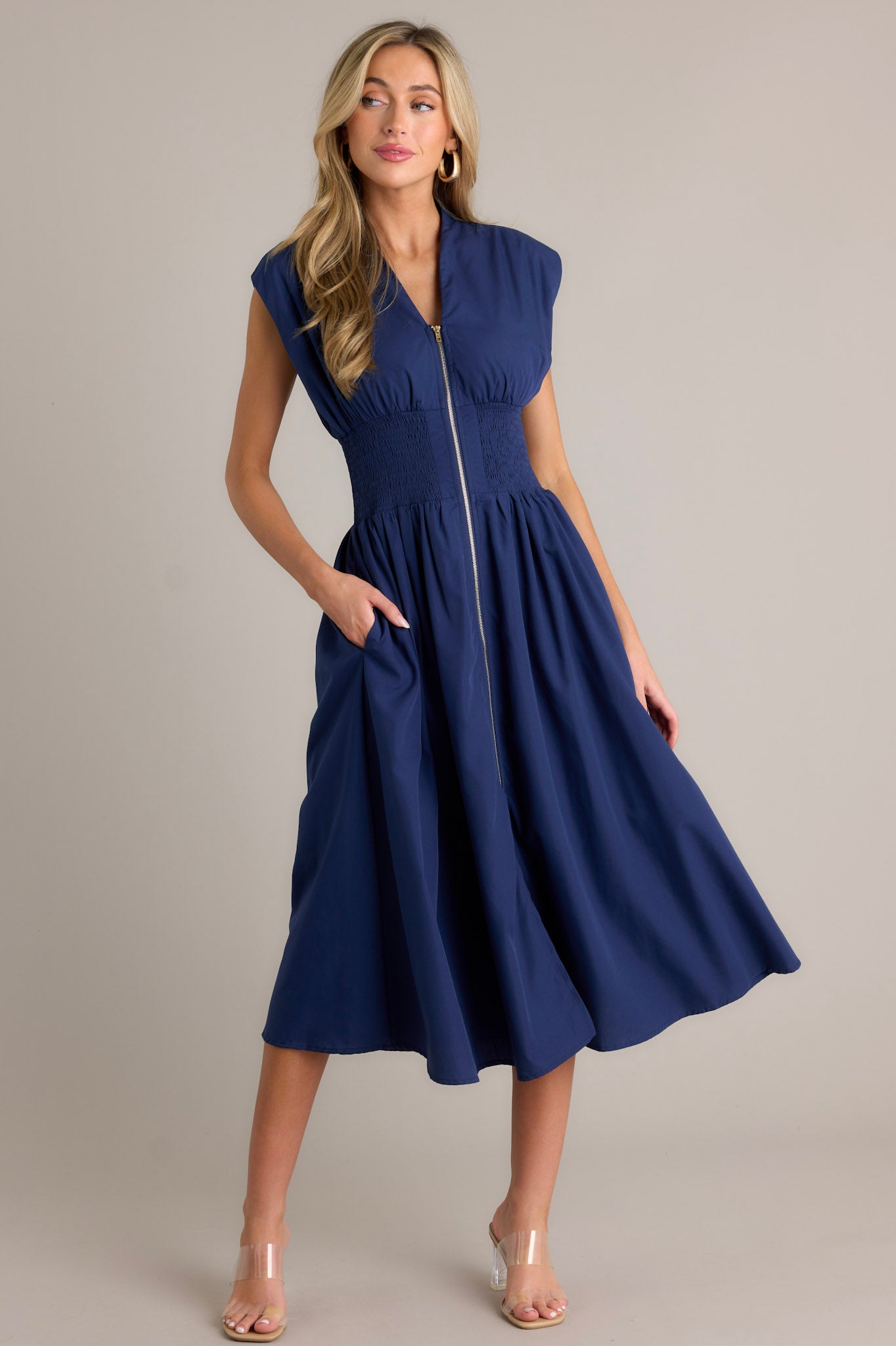 This navy midi dress features a v-neckline, padded shoulders, a full zipper front, a fully smocked waist, functional hip pockets, and a front slit.