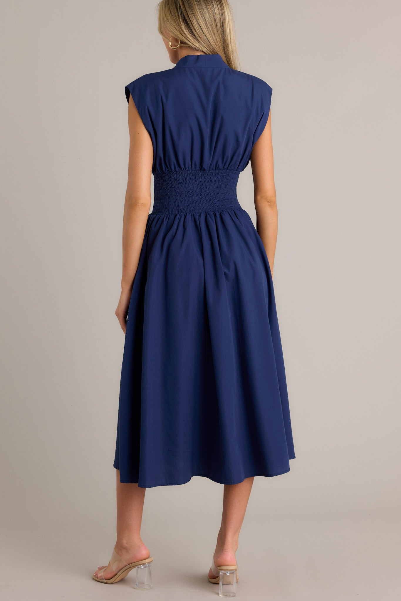 Back view of a navy blue dress with a V-neckline, cap sleeves, a zippered front, and a cinched waist with shirred detailing, featuring a flowing midi-length skirt, highlighting its elegant and stylish design.