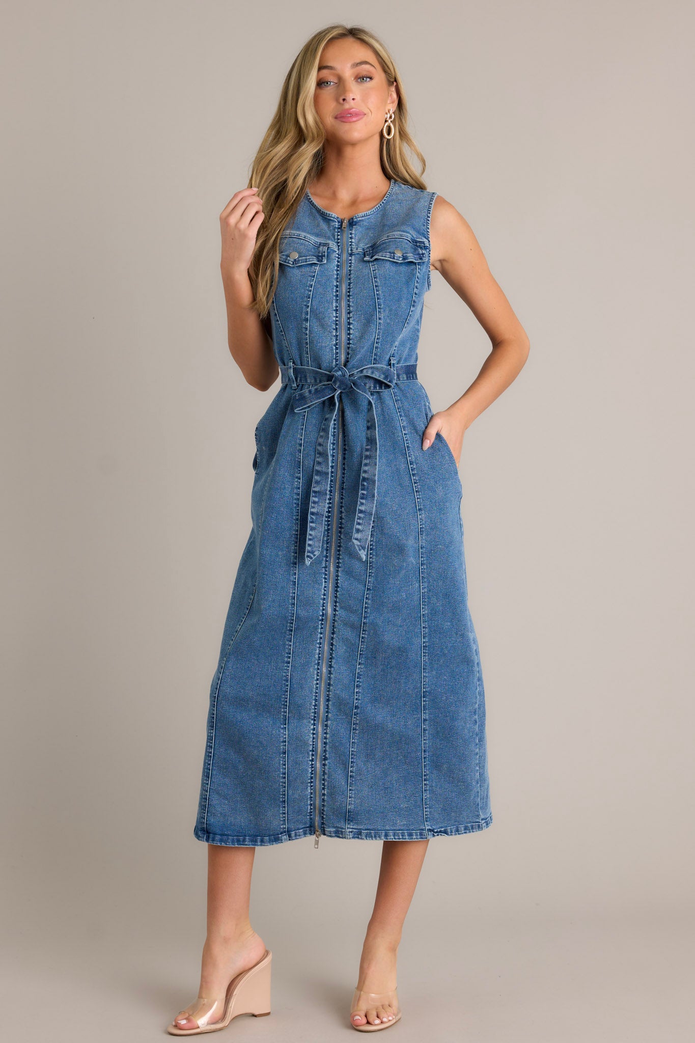 Front view of a denim dress featuring a rounded neckline, a functional front zipper, faux chest pockets, functional hip pockets, a self-tie denim drawstring belt, and a sleeveless design.