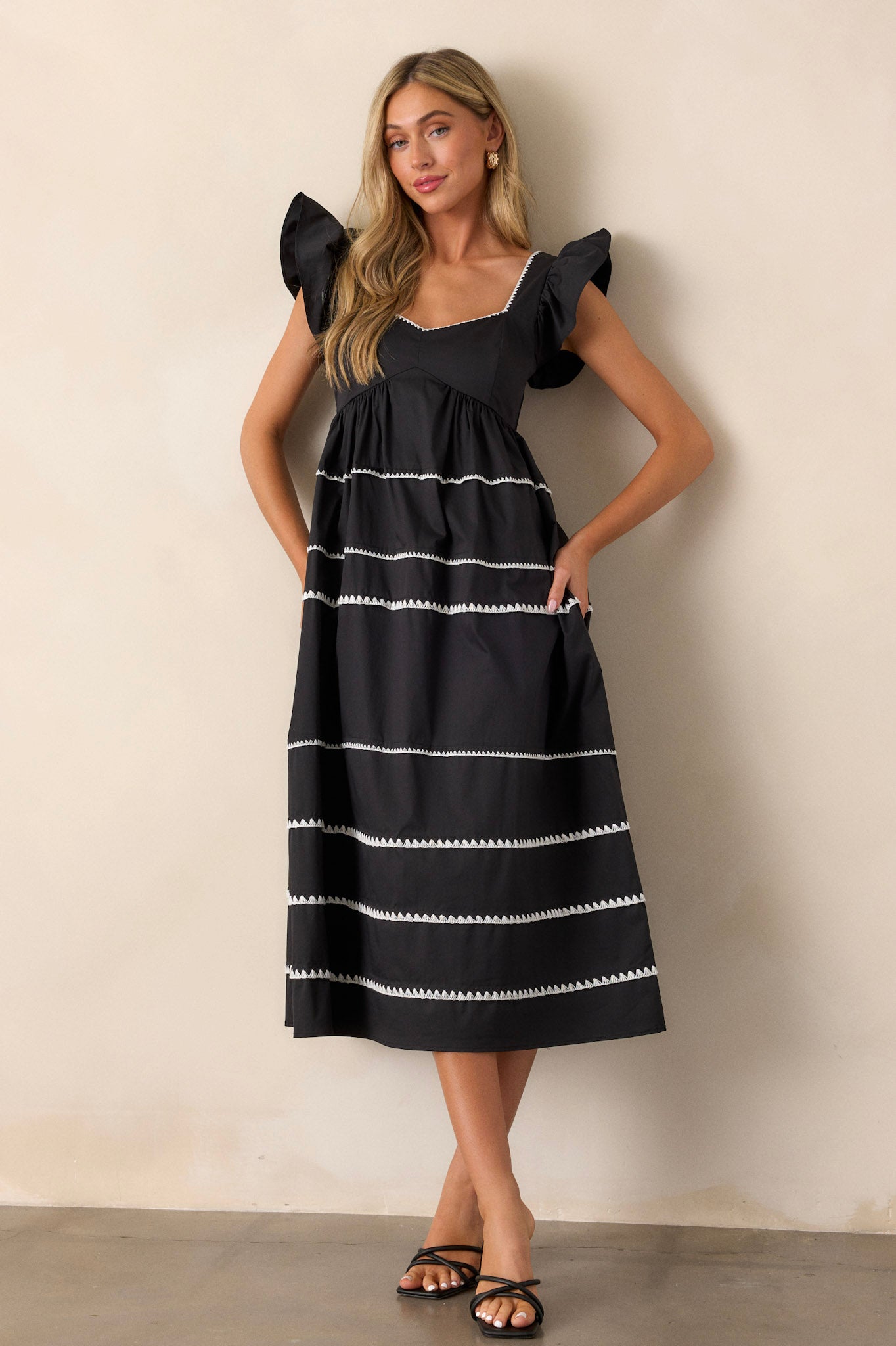 Front view of a black dress featuring ruffle cap sleeves, a smocked back, multiple tiers, two functional pockets, and ric-rac detailing.