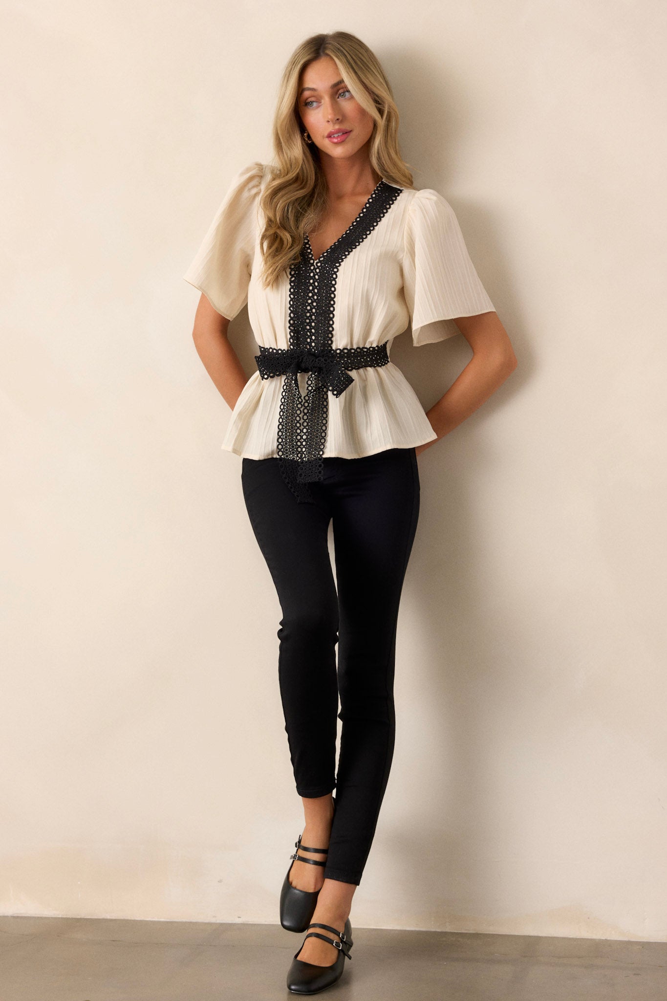 Front view of an ivory lace top featuring a v-neckline, black circle lace detailing on the neckline and down the bodice, a faux lace waist belt, an elastic waist insert, and short sleeves.