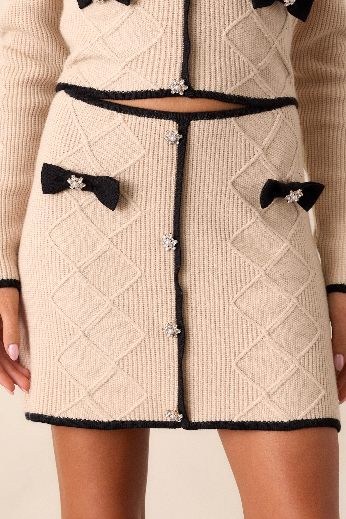 Close-up view of this beige mini skirt that features black bow embellishments, pearl and crystal faux buttons up the front, black trim, and a soft knit fabric