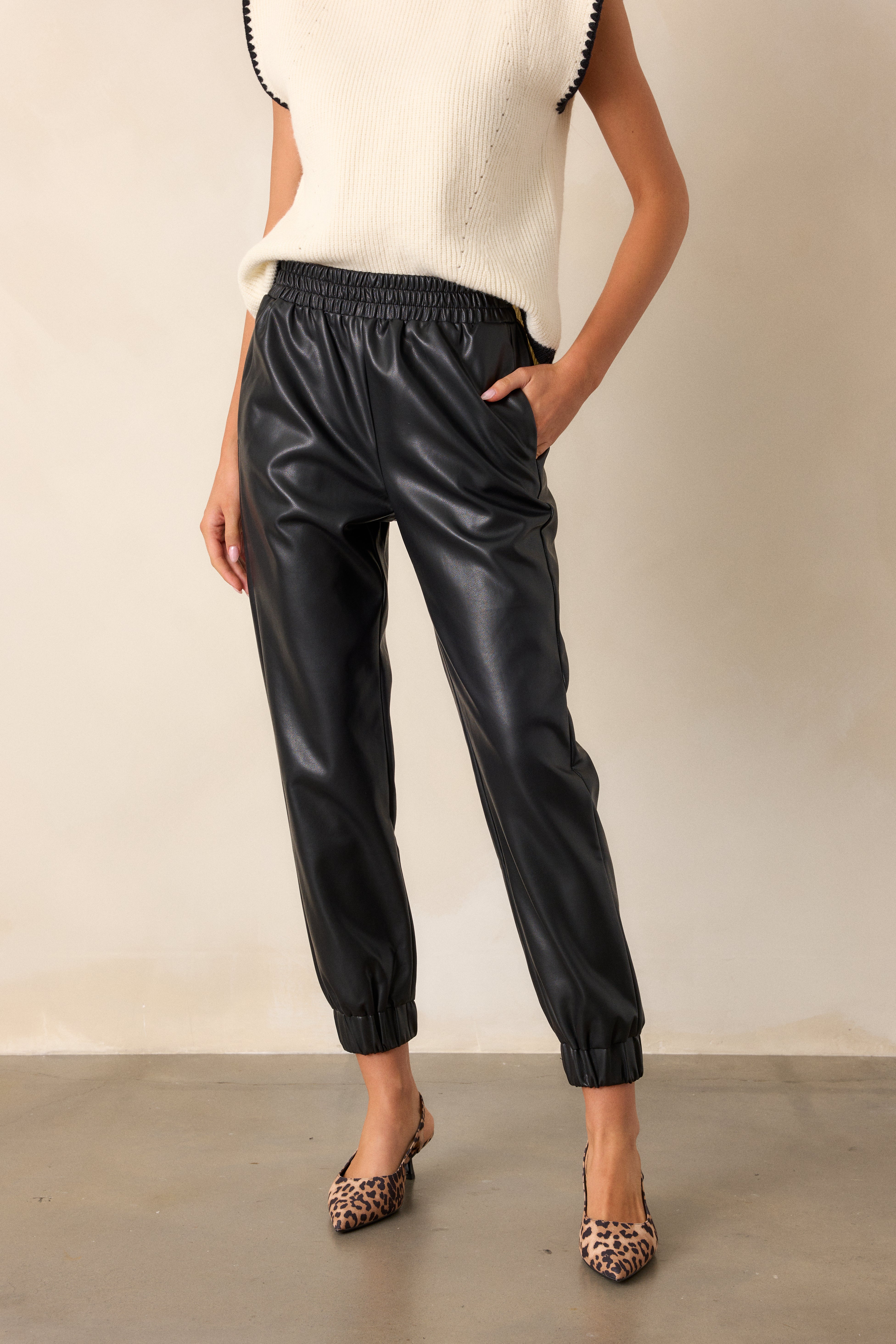 Full length view of black pants featuring an elastic waistband, functional hip and back side pockets, elastic cuffed ankles, and a faux leather material