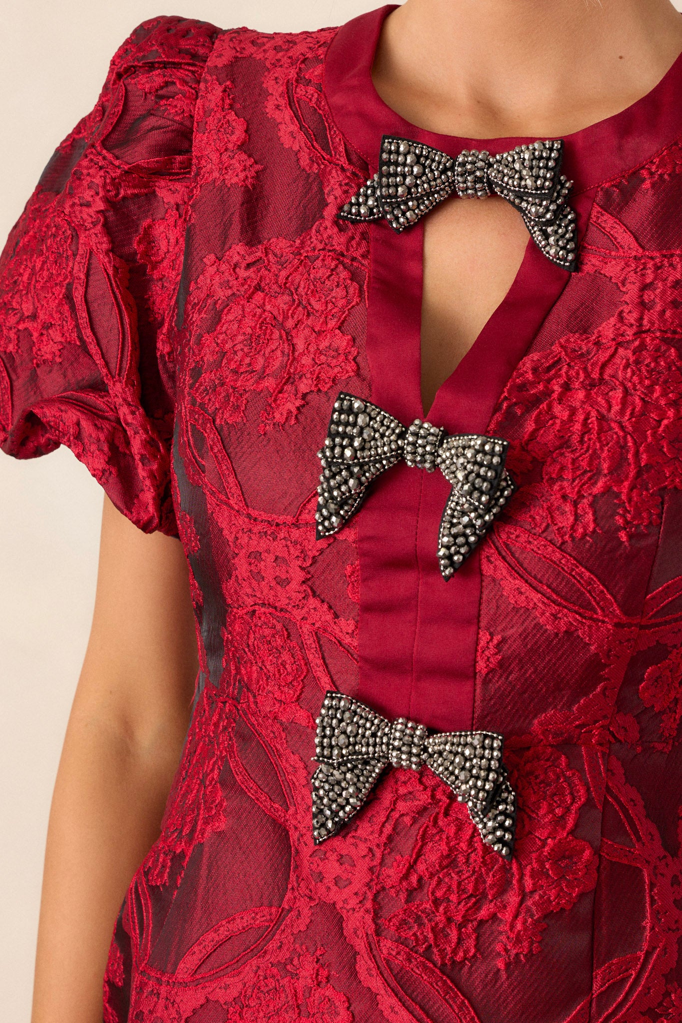 Close-up of the berry red dress focusing on the triangular cutout at the neckline, the three bows down the bodice, and the texture of the short elastic cuffed puff sleeves.