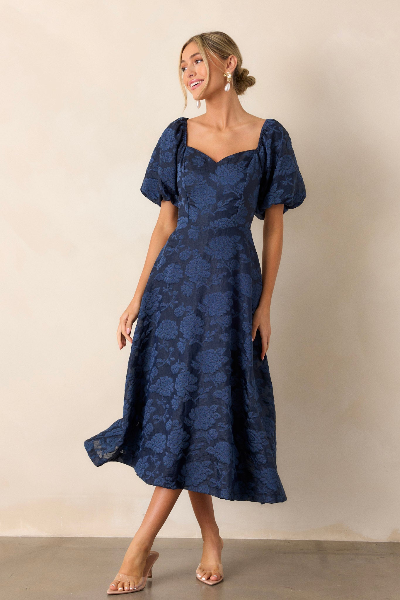 Front angled view of the navy floral dress featuring a Queen Anne neckline, a fitted bodice, a discrete back zipper, floral design, and elastic cuffed puff sleeves