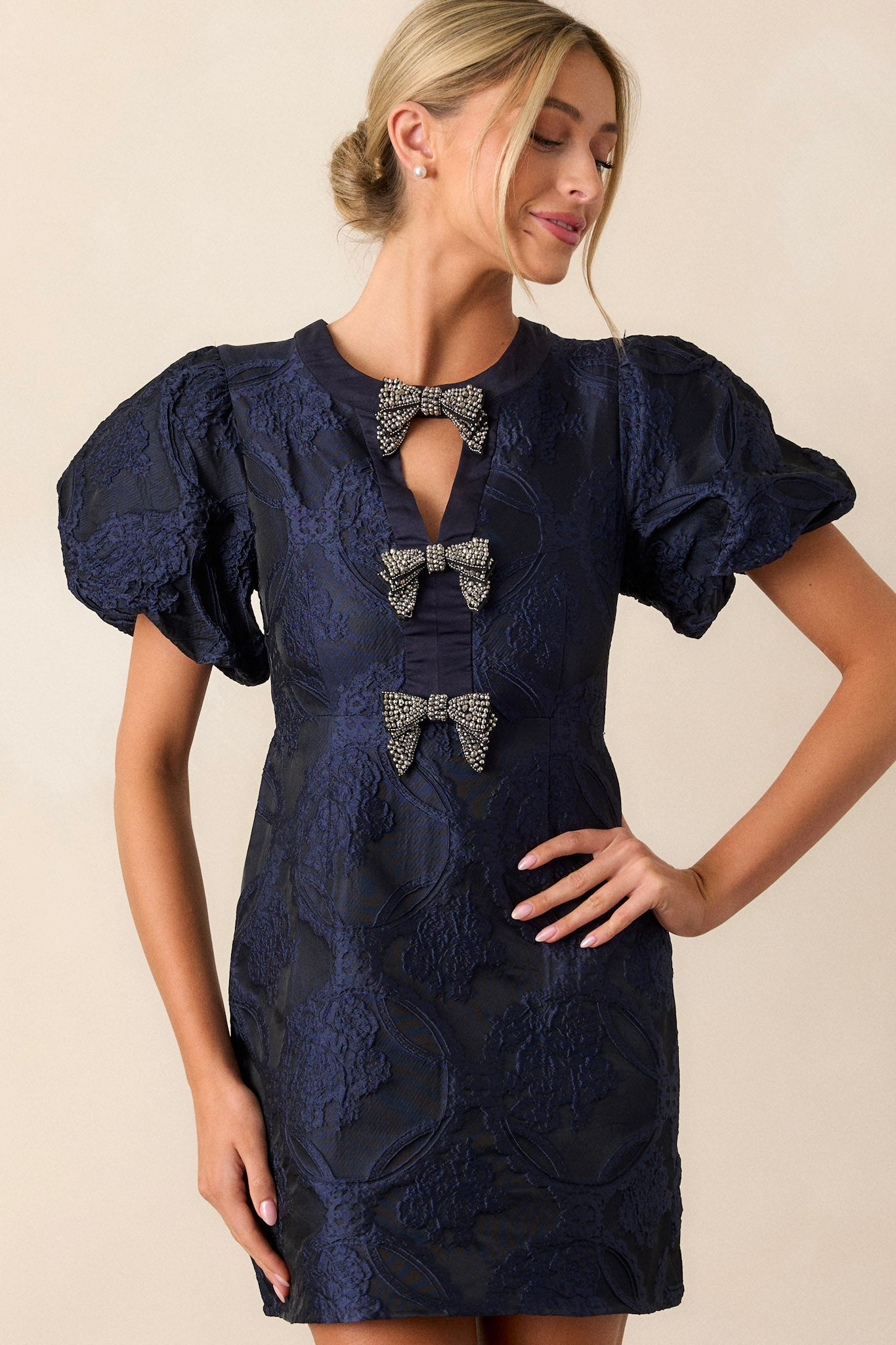 Front angled view of this navy dress featuring a high neckline with a triangular cutout, three bows down the bodice, a discrete back zipper, and short elastic cuffed puff sleeves