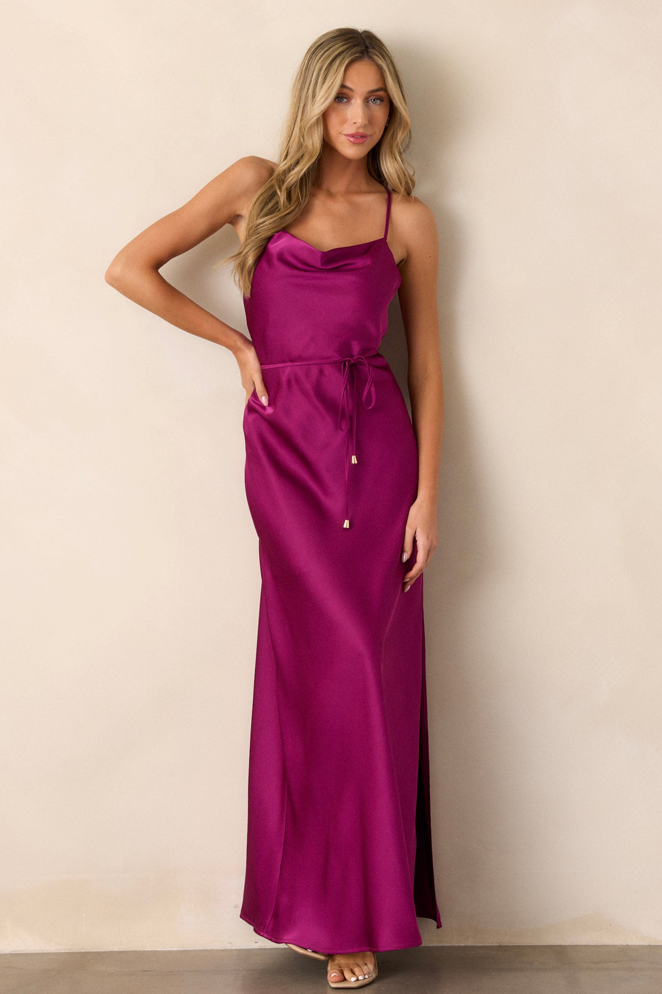 Full-length view of a model wearing the magenta dress, emphasizing the cowl neckline, adjustable straps, the self-tie belt with gold hardware, the knee-length slit on the left side, and the silky satin material.