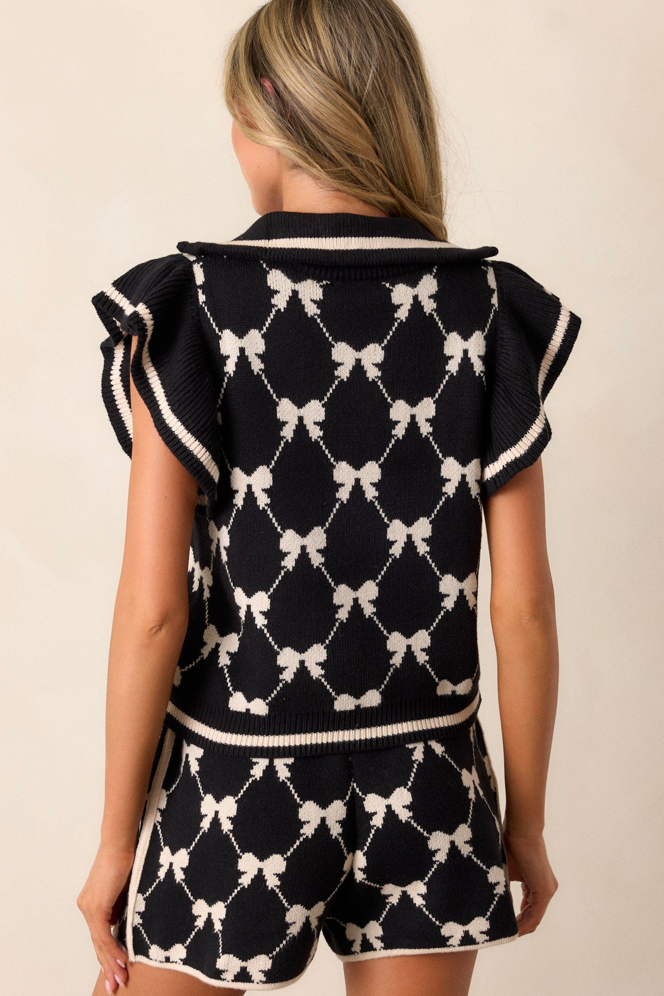 Back view of this black top that features a functional quarter zip-up front, a collared neckline, a white bow print, flutter sleeves, and a soft knit fabric.