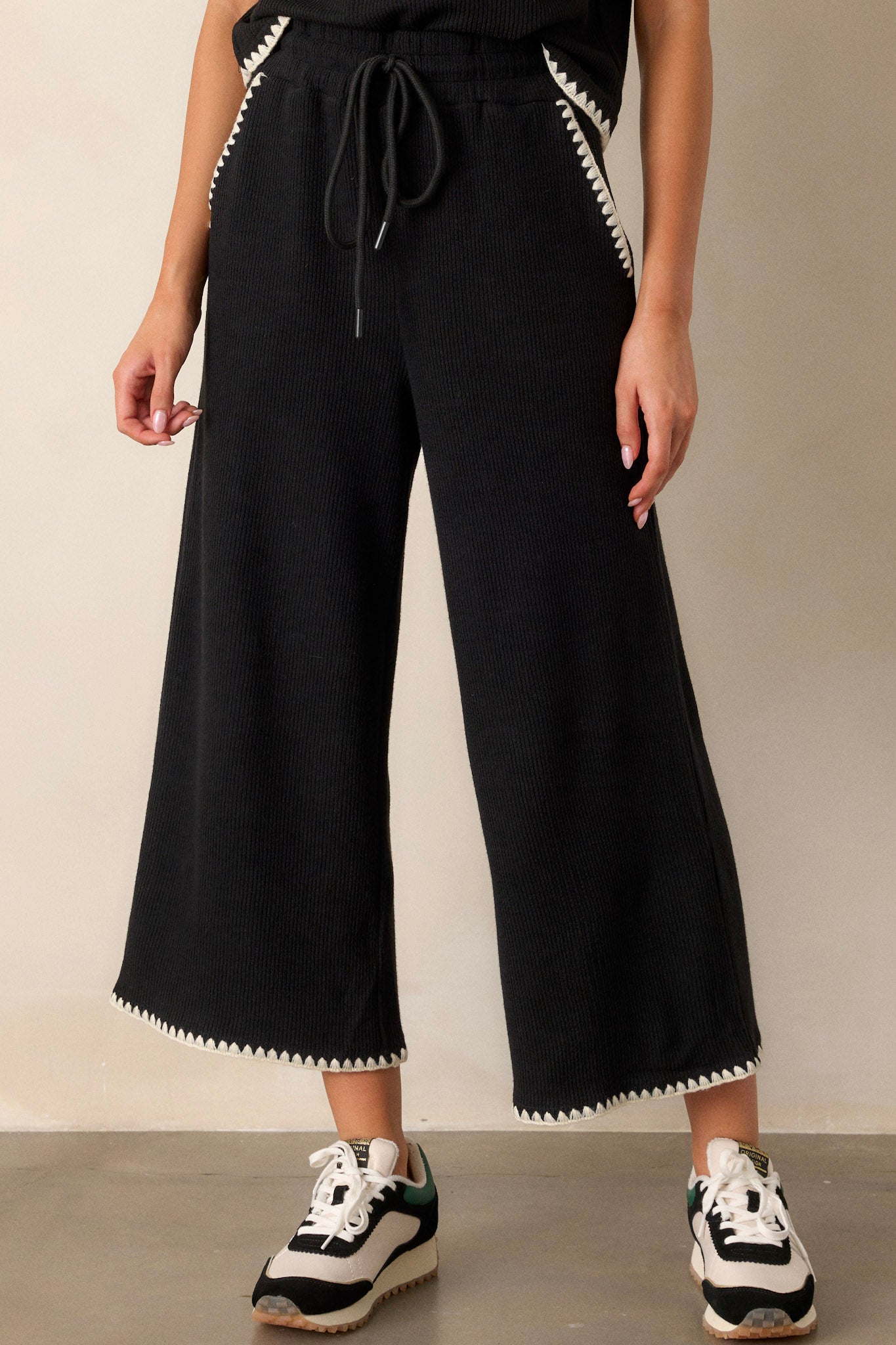 Close-up of these black pants that feature an elastic waistband, an optional waist drawstring, an ivory ruffle trim, functional hip pockets, a soft stitched material and a wide leg