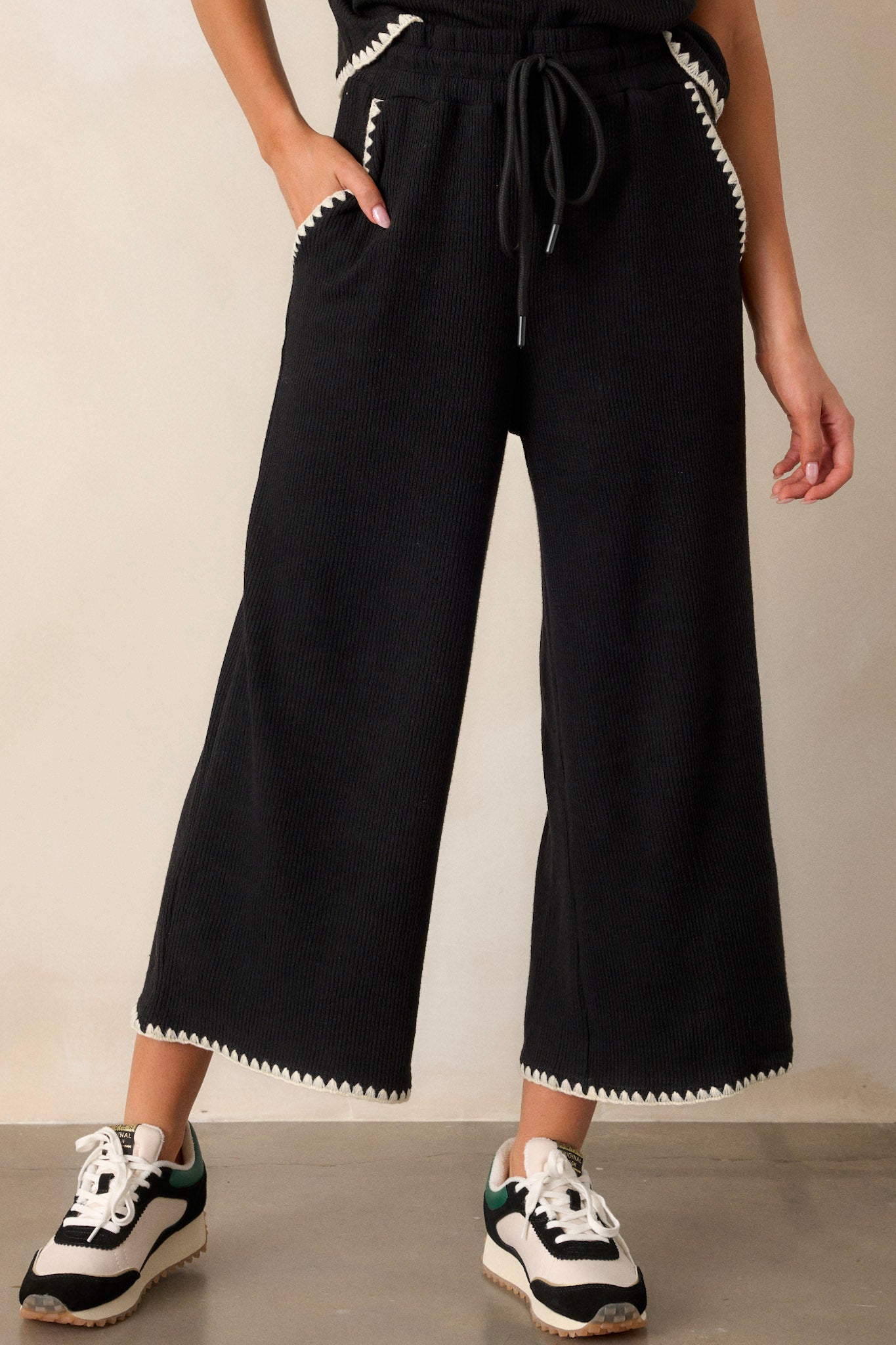 Front view of these black pants that feature an elastic waistband, an optional waist drawstring, an ivory ruffle trim, functional hip pockets, a soft stitched material and a wide leg