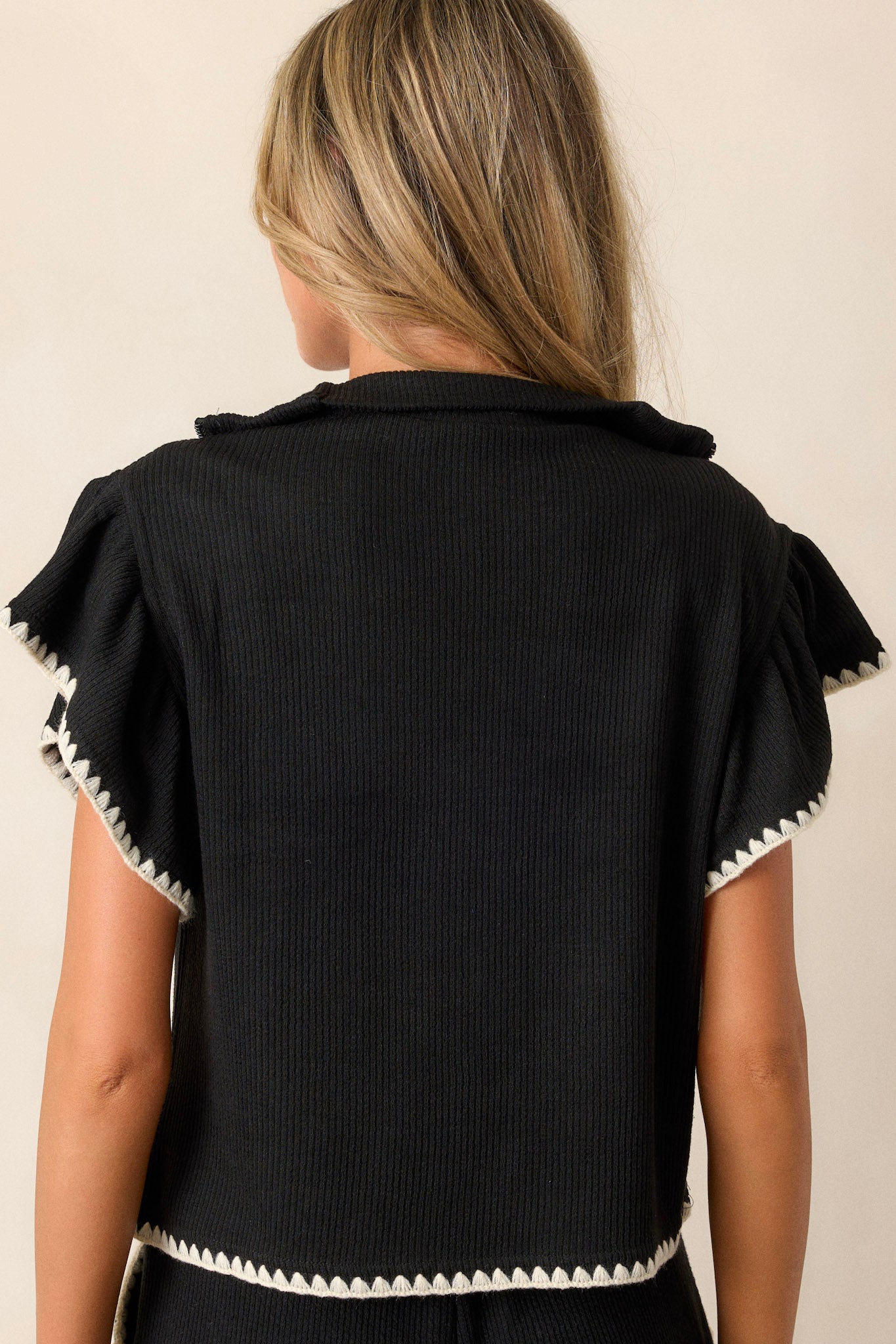 Back view of this black sweater that features a collared quarter zip neckline, a relaxed fit, a soft knit material, flutter sleeves, and ivory blanket stitch detailing.