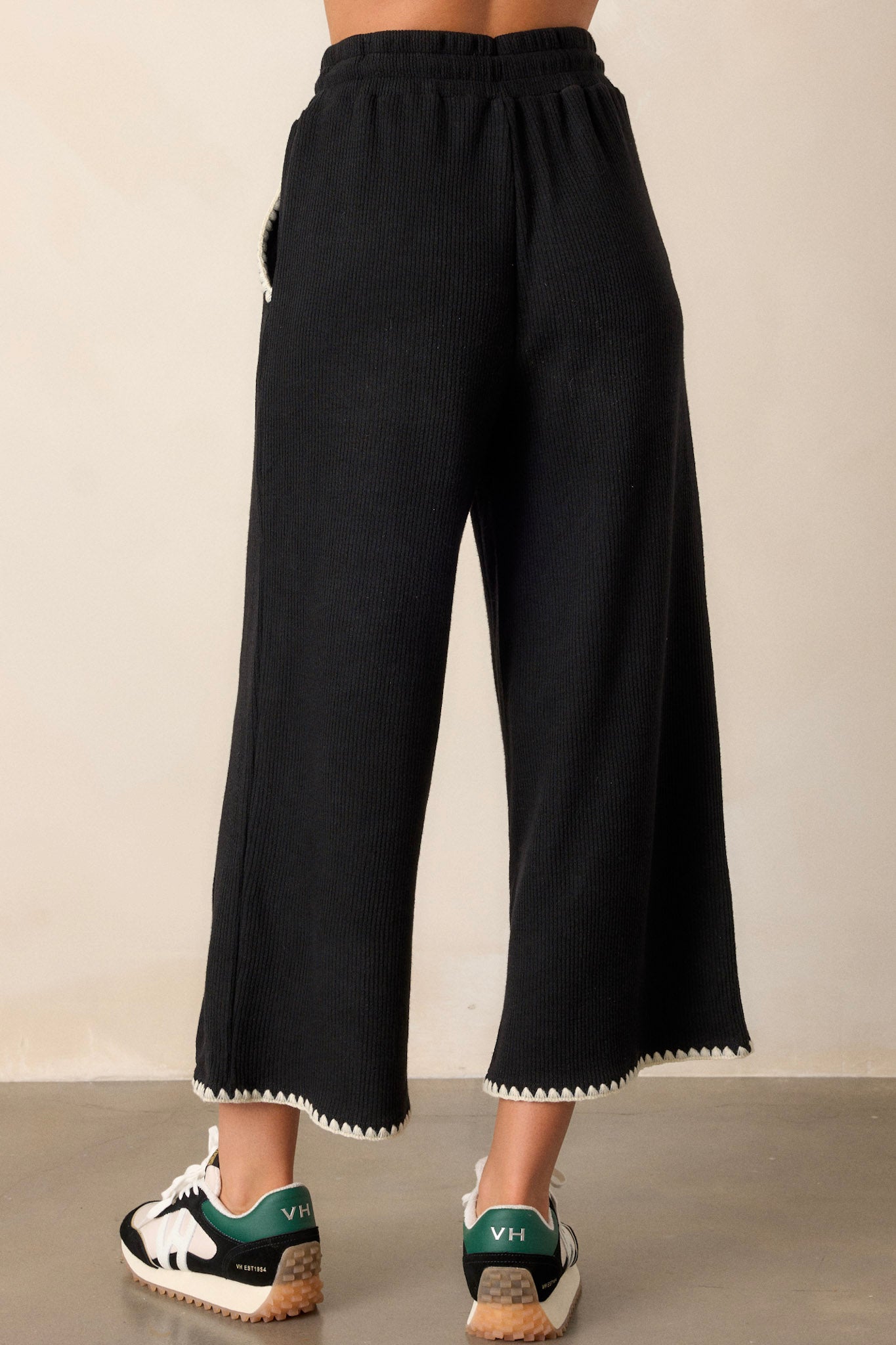 Back of these black pants that feature an elastic waistband, an optional waist drawstring, an ivory ruffle trim, functional hip pockets, a soft stitched material and a wide leg