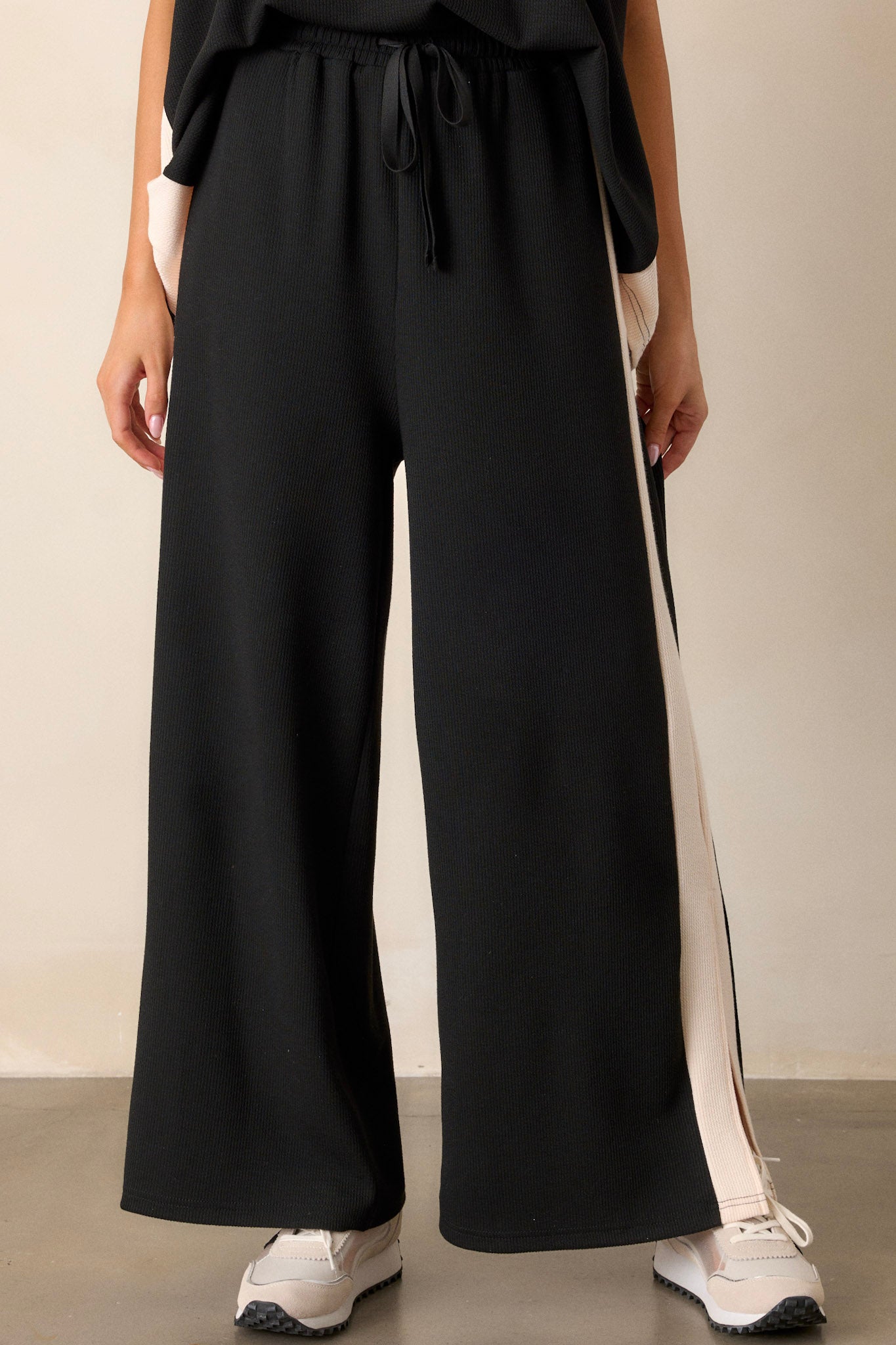 Front view of black and ivory pants featuring an elastic waistband, a high waisted design, a waist drawstring, soft ribbed texture, functional hip pockets, white trim down the sides of the legs, and high slits in the legs