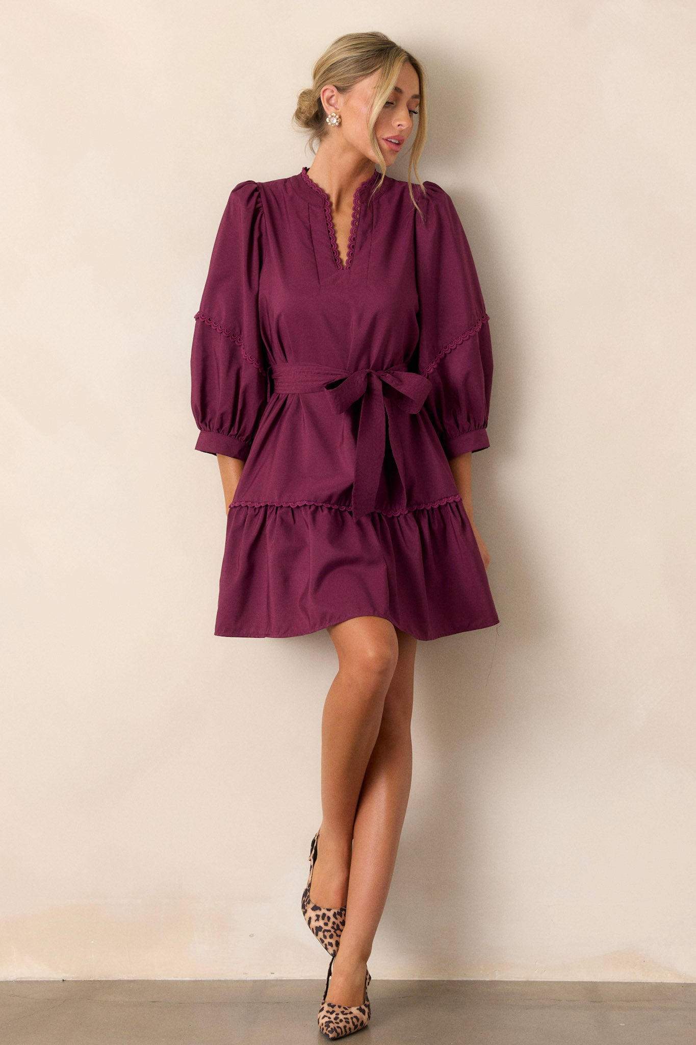 Front angled view of the plum mini dress showcasing the split neckline with scalloped detailing, the self-tie drawstring belt, functional hip pockets, the tiered design, and the bishop sleeves with scalloped detailing