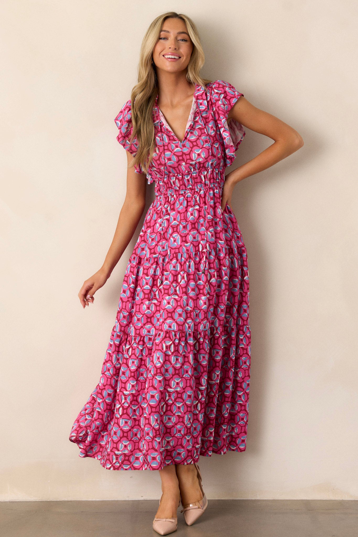 Front view of a pink maxi dress featuring a ruffled v-neckline, a self-tie drawstring neckline, elastic waistbands, functional hip pockets, a tiered design, and short ruffle sleeves.