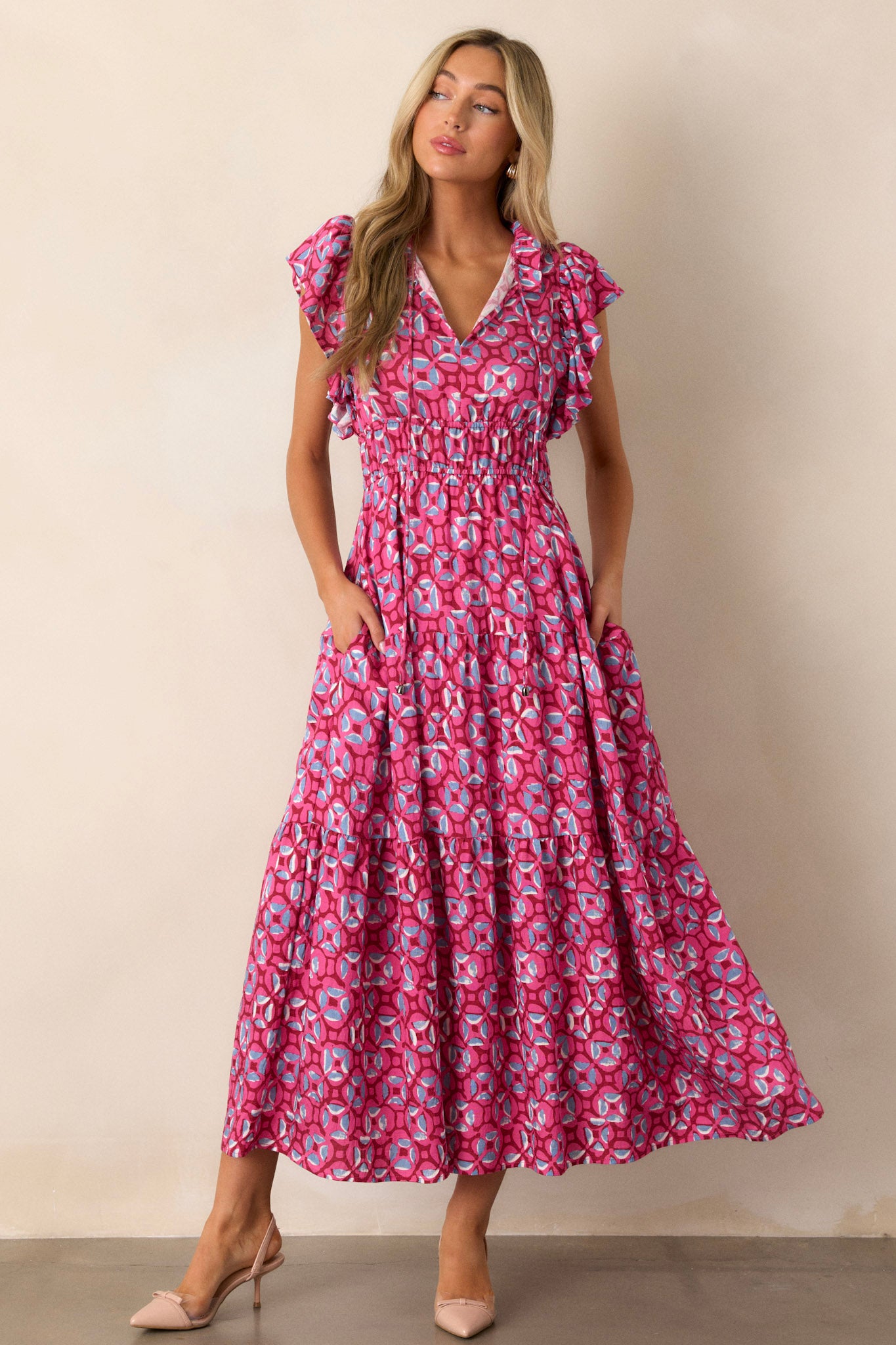 Front angled view of the pink maxi dress featuring a ruffled v-neckline, self-tie drawstring neckline, elastic waistbands, functional hip pockets, a tiered design, and short ruffle sleeves