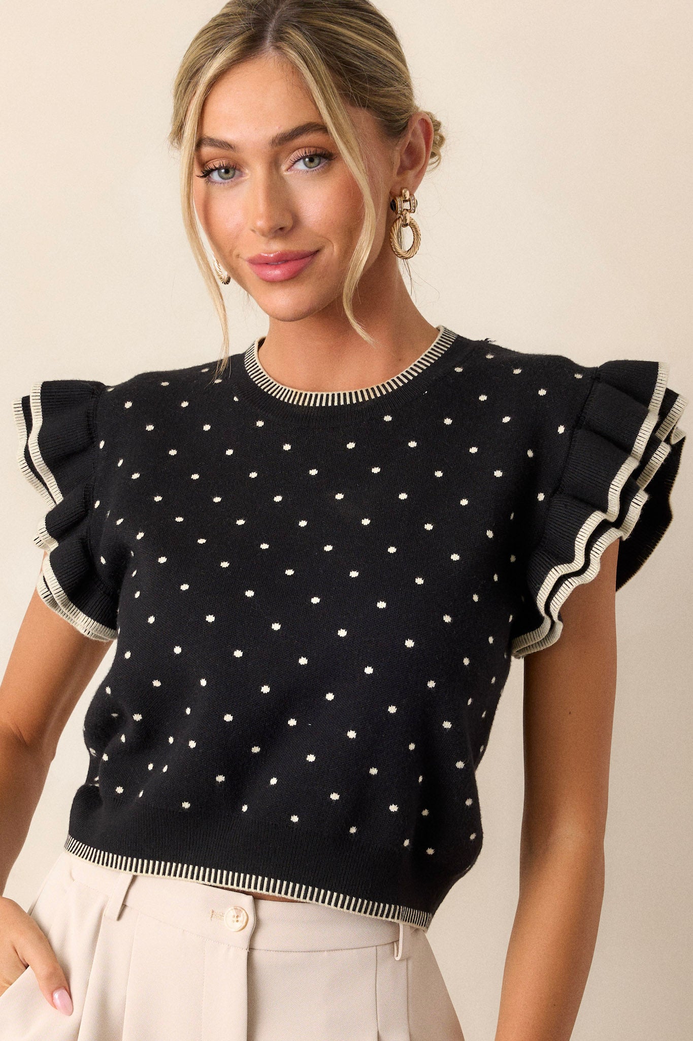 This black sweater features an ivory polka dot print, blanket stitching along bottom hem, blanket stitching along the crew neckline, flutter sleeves, and a knit fabric.