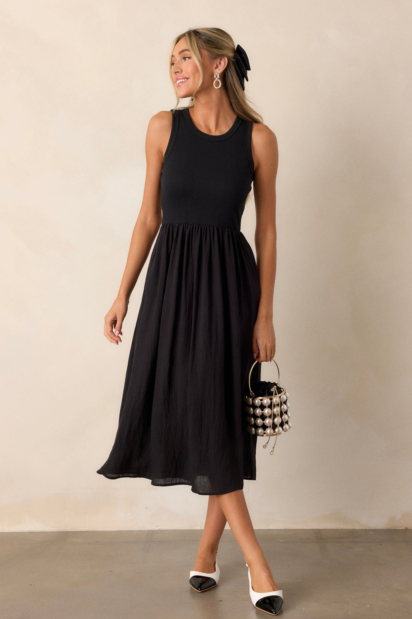 This sleeveless black dress features a crew neckline, a stretchy ribbed bust, a cinched waist, and flowing mid-length skirt.