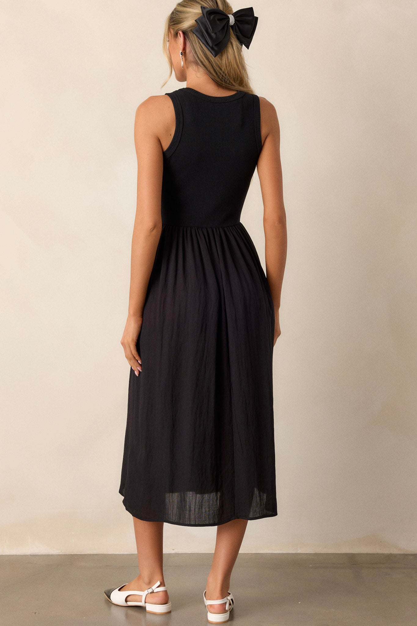 Back view of this sleeveless black dress that features a crew neckline, a stretchy ribbed bust, a cinched waist, and flowing mid-length skirt.