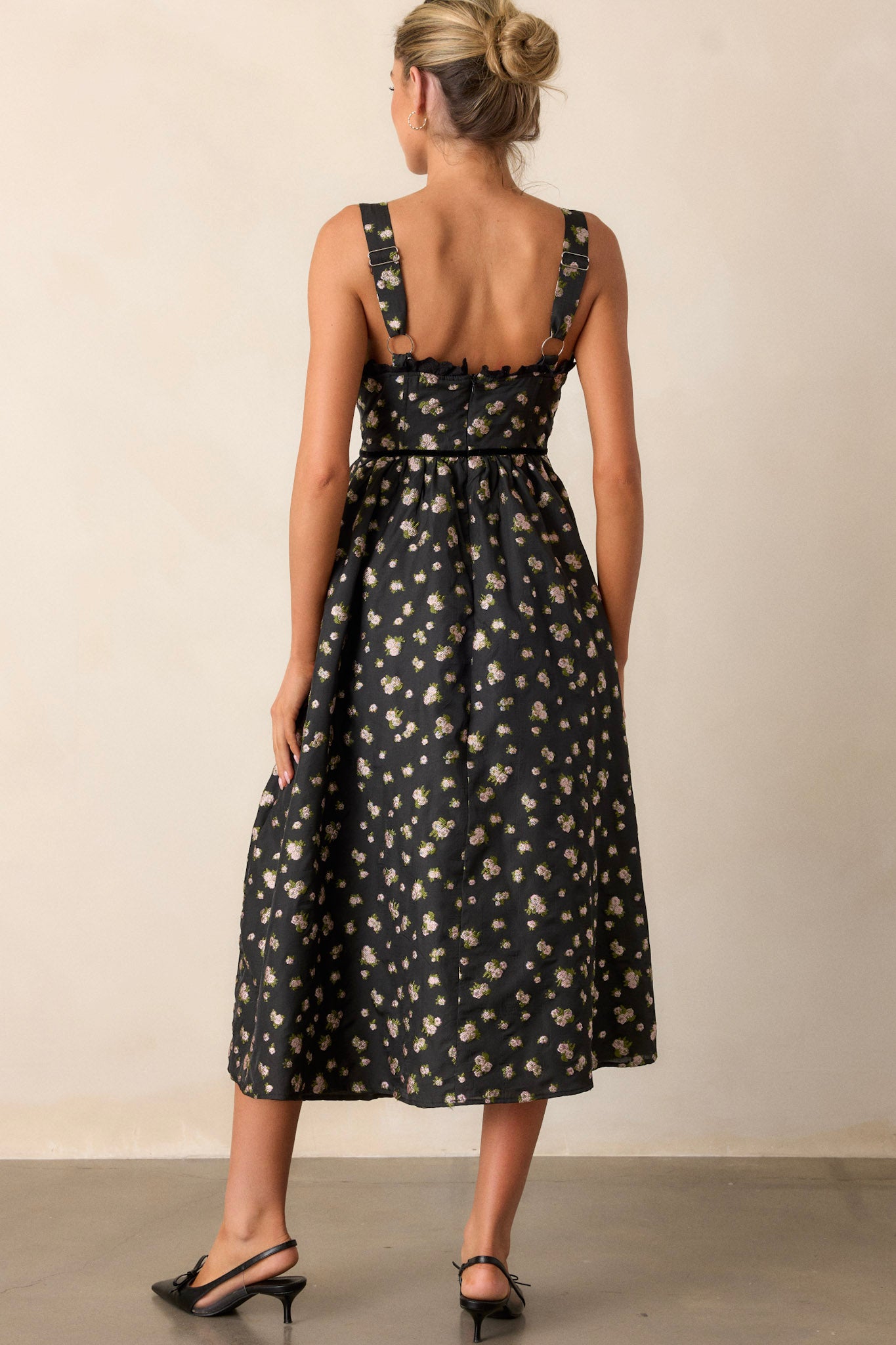 Back view of this black dress that features a back functional zipper, a corset style top, adjustable straps, a flowing skirt, an ivory floral print, and lace detailing.