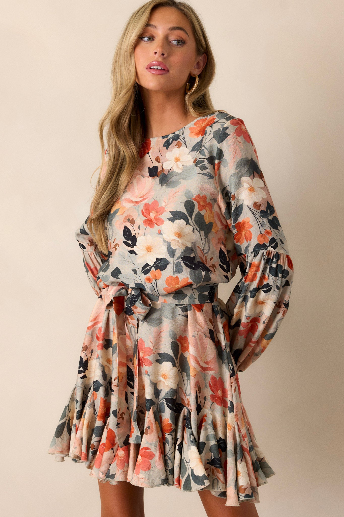 Front angled view of the green floral dress featuring a high neckline, an orange and white floral print, a self-tie waist belt, a ruffled hem, a button loop back closure, and elastic cuffed long sleeves