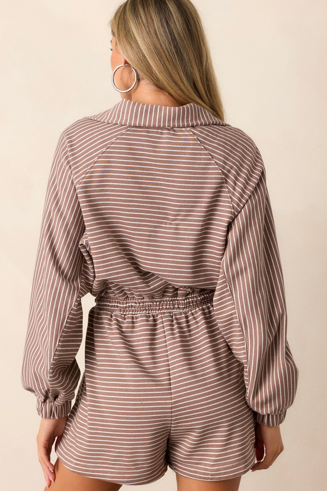 Back view of this mocha stripe romper that features a collared neckline, a functional front zipper, a brown & ivory striped design, an elastic waistband, functional hip pockets, a waist drawstring and long sleeves.