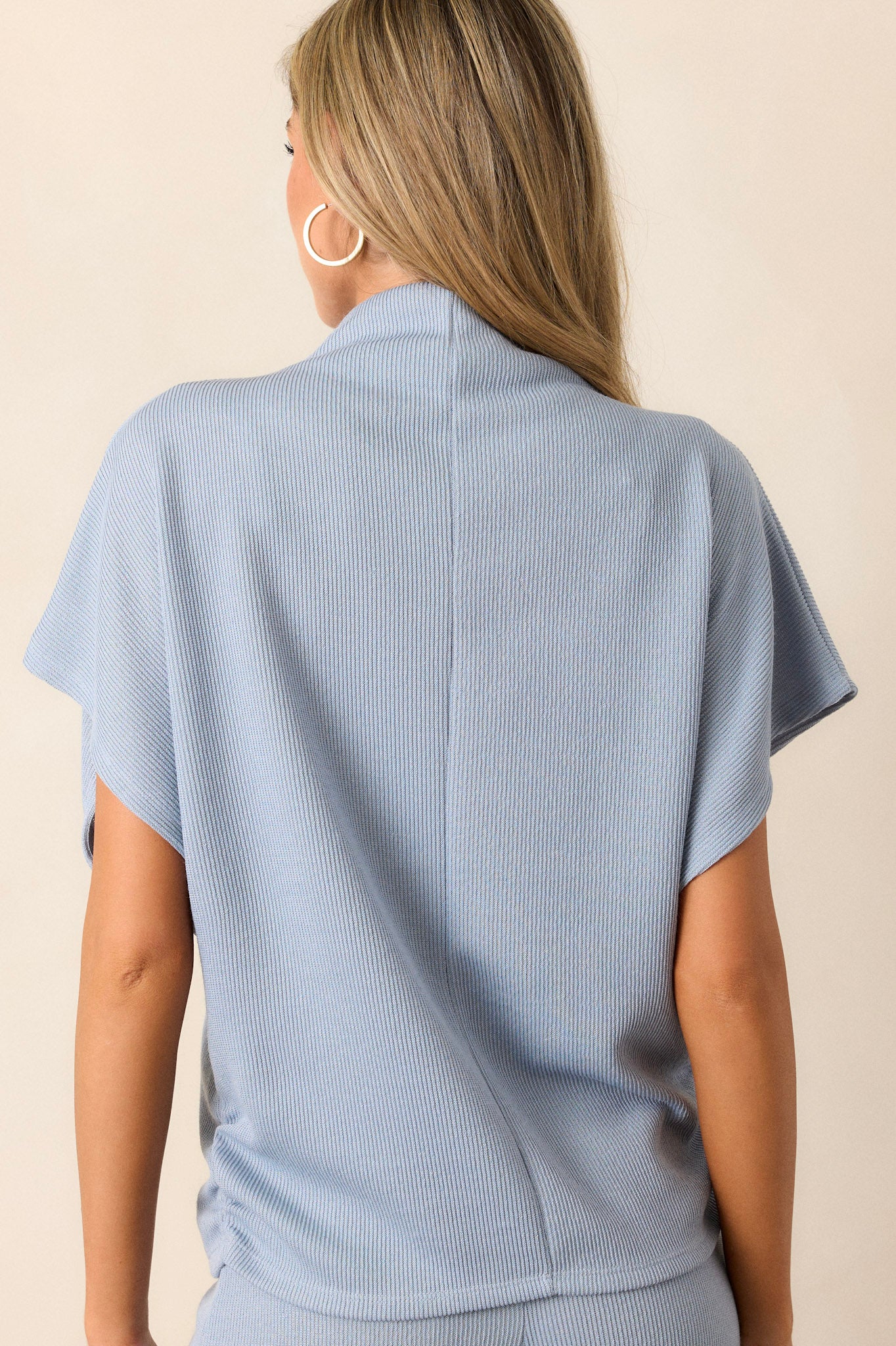 back view of this blue top that features a turtleneck, a knit towel like texture on the outside, a soft smooth inside, cinching in the hips and short flowy sleeves.