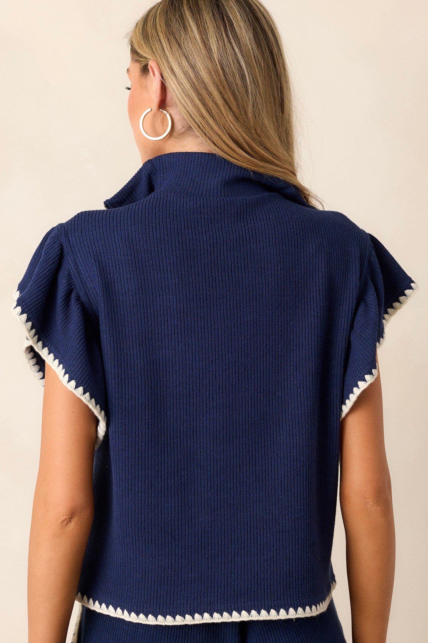 Back view of this navy sweater that features a collared quarter zip neckline, a relaxed fit, a soft knit material, flutter sleeves, and ivory blanket stitch detailing.