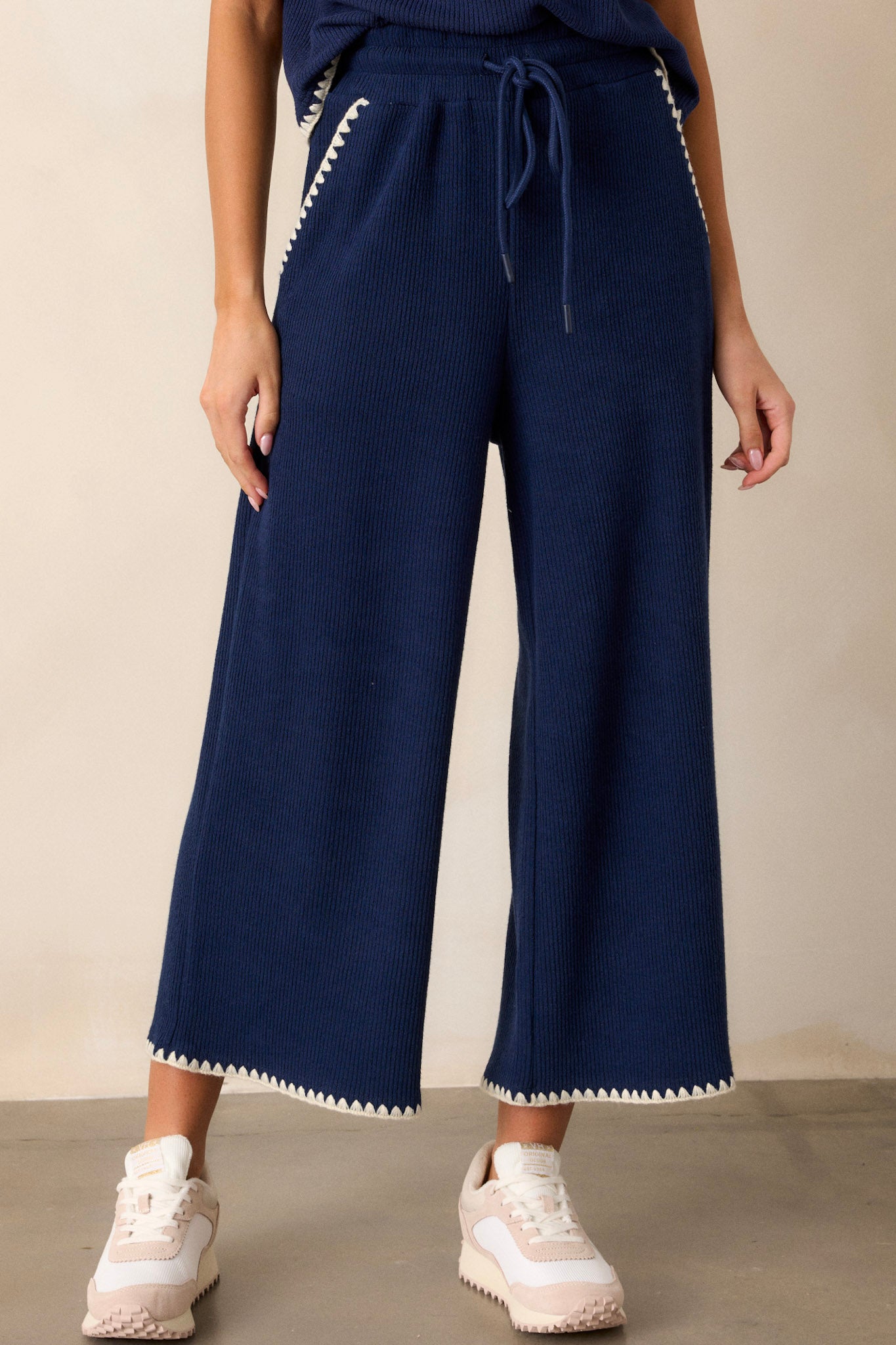 Front view of these navy pants that feature an elastic waistband, an adjustable drawstring, ivory blanket stitch detailing, functional hip pockets, a soft knit material, and a wide leg.