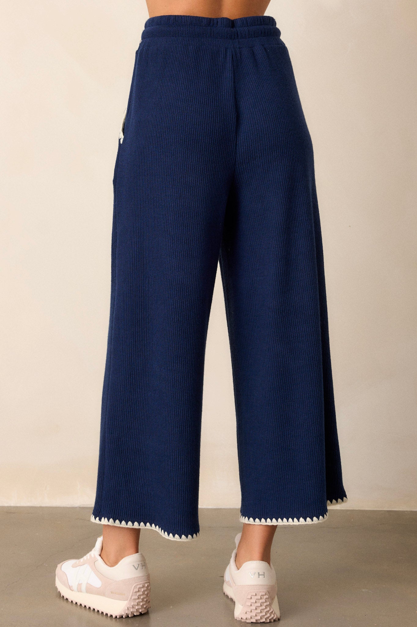 Back view of these navy pants that feature an elastic waistband, an adjustable drawstring, ivory blanket stitch detailing, functional hip pockets, a soft knit material, and a wide leg.